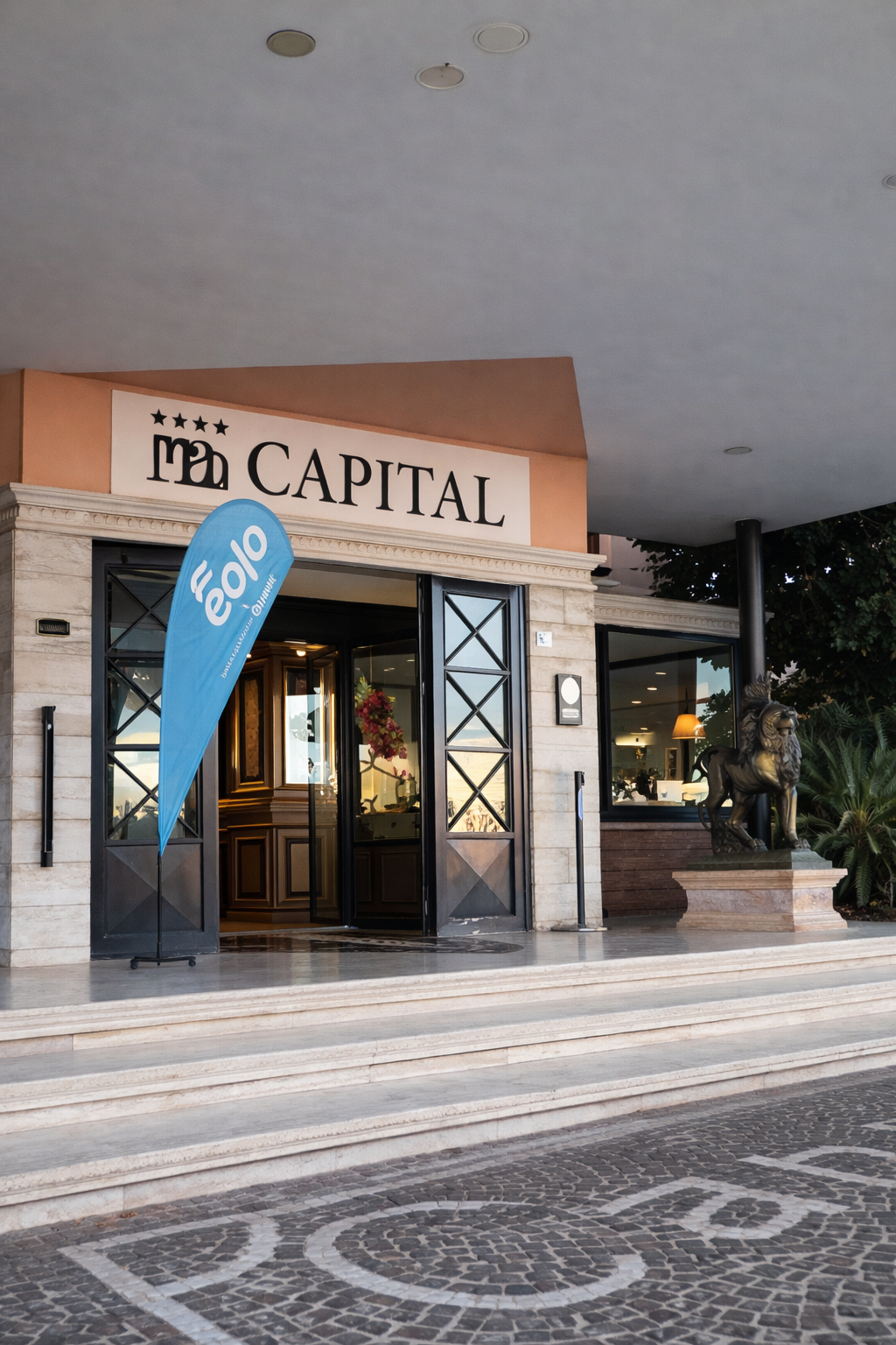 Ingresso dell'Hotel Capital, con porte aperte, cartello blu ed esterno beige.