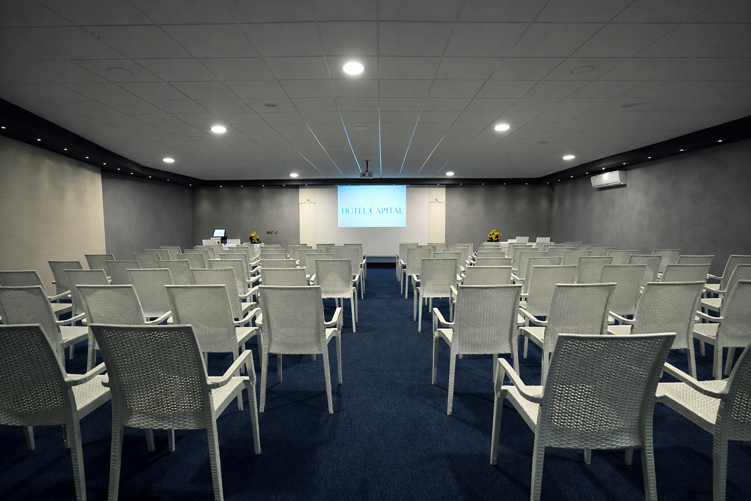 Sala conferenze vuota con file di sedie bianche di fronte a uno schermo per proiettore su un tappeto blu scuro.