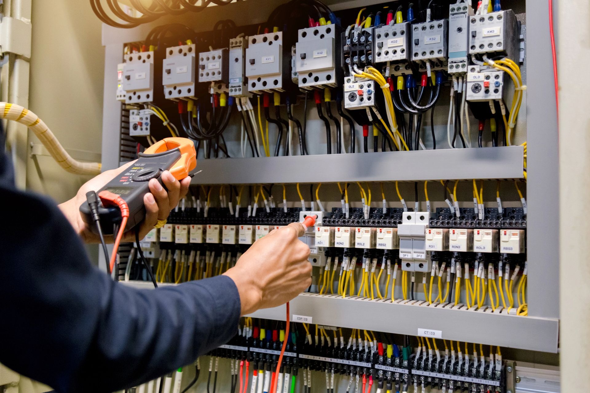 Elettricista che utilizza un multimetro per ispezionare il cablaggio in un quadro elettrico, con vari fili e componenti visibili.