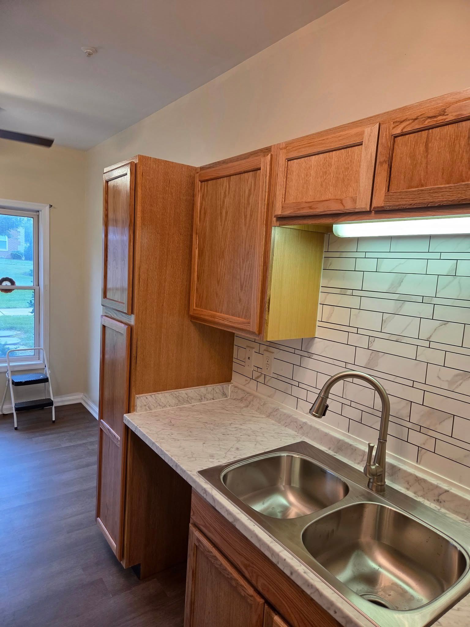 Small kitchen with wood cabinets, double sink, marble counters, and a window at the end of the room.