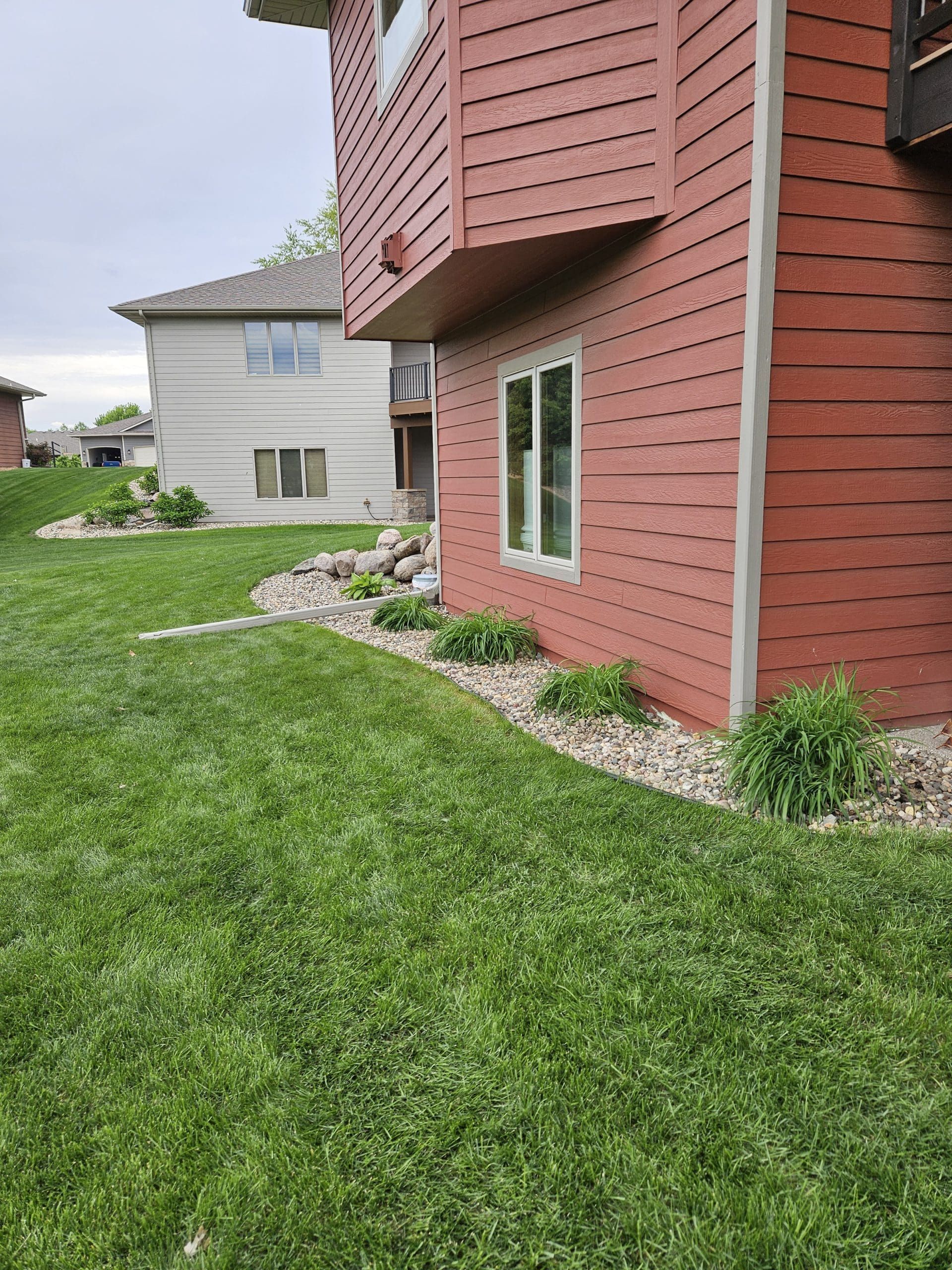 Red-sided house with green lawn and rock border, another building in the background.