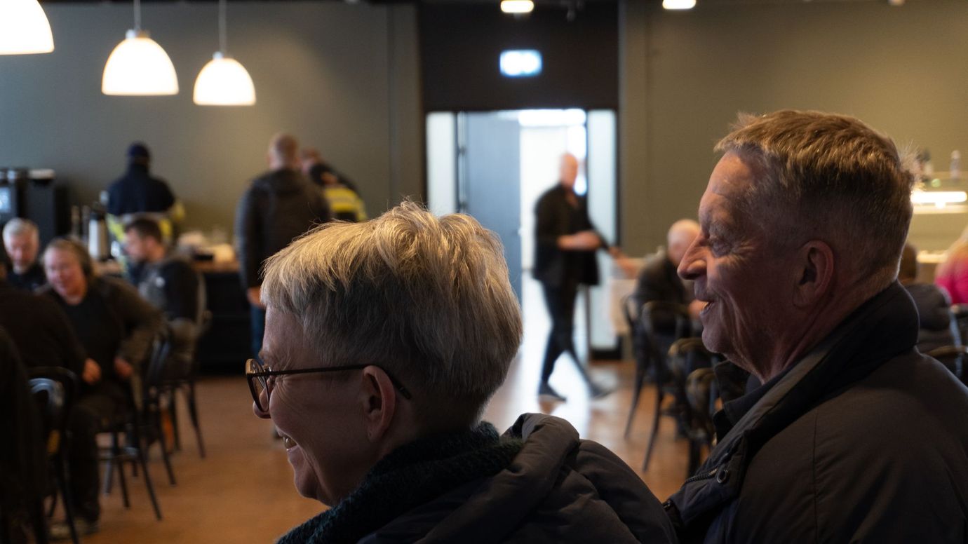 Audience seated in a dim café or event space, listening toward a bright doorway and speaker.
