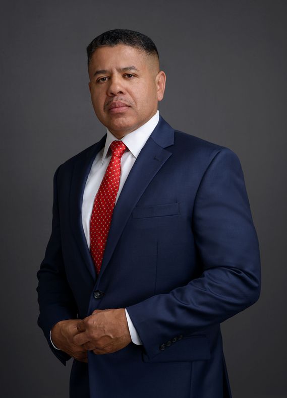 A man in a blue suit and red tie is standing with his hands folded.
