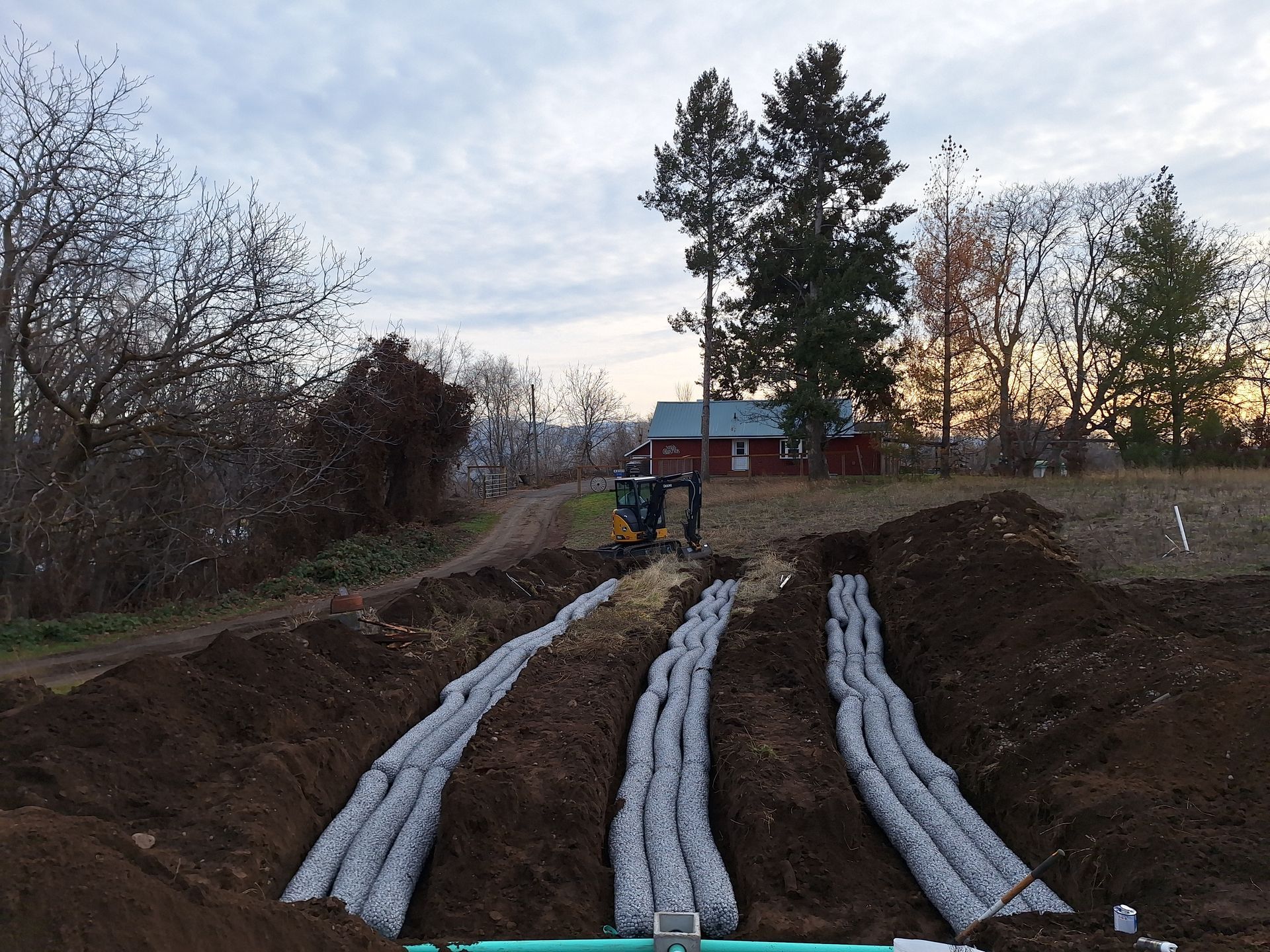 Construction site with trenches, gravel-filled tubes, and a small excavator. Dirt mounds, trees, and a house in the background.