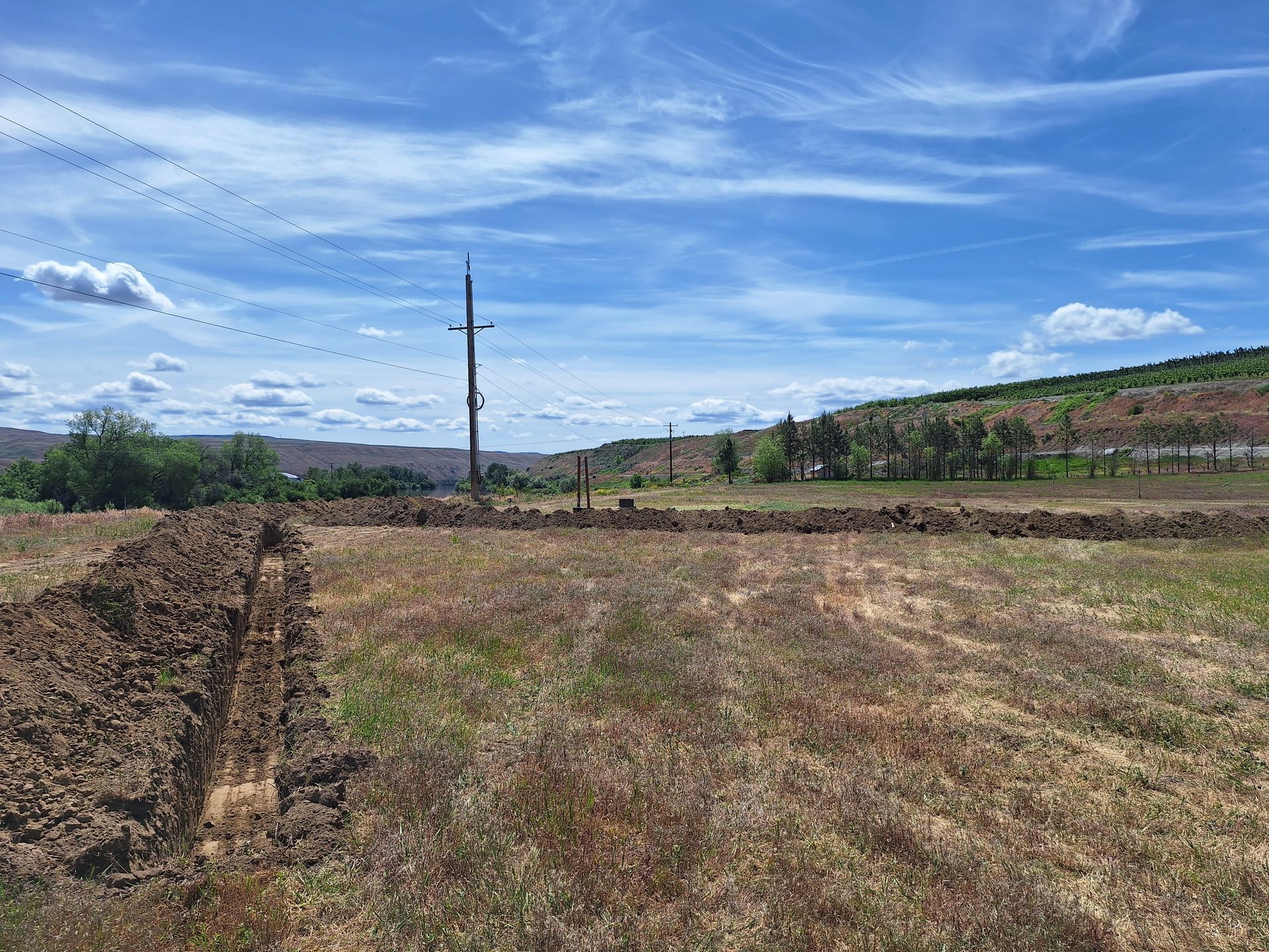 Field with brown dirt trench, power lines, green grass, and blue sky with clouds.