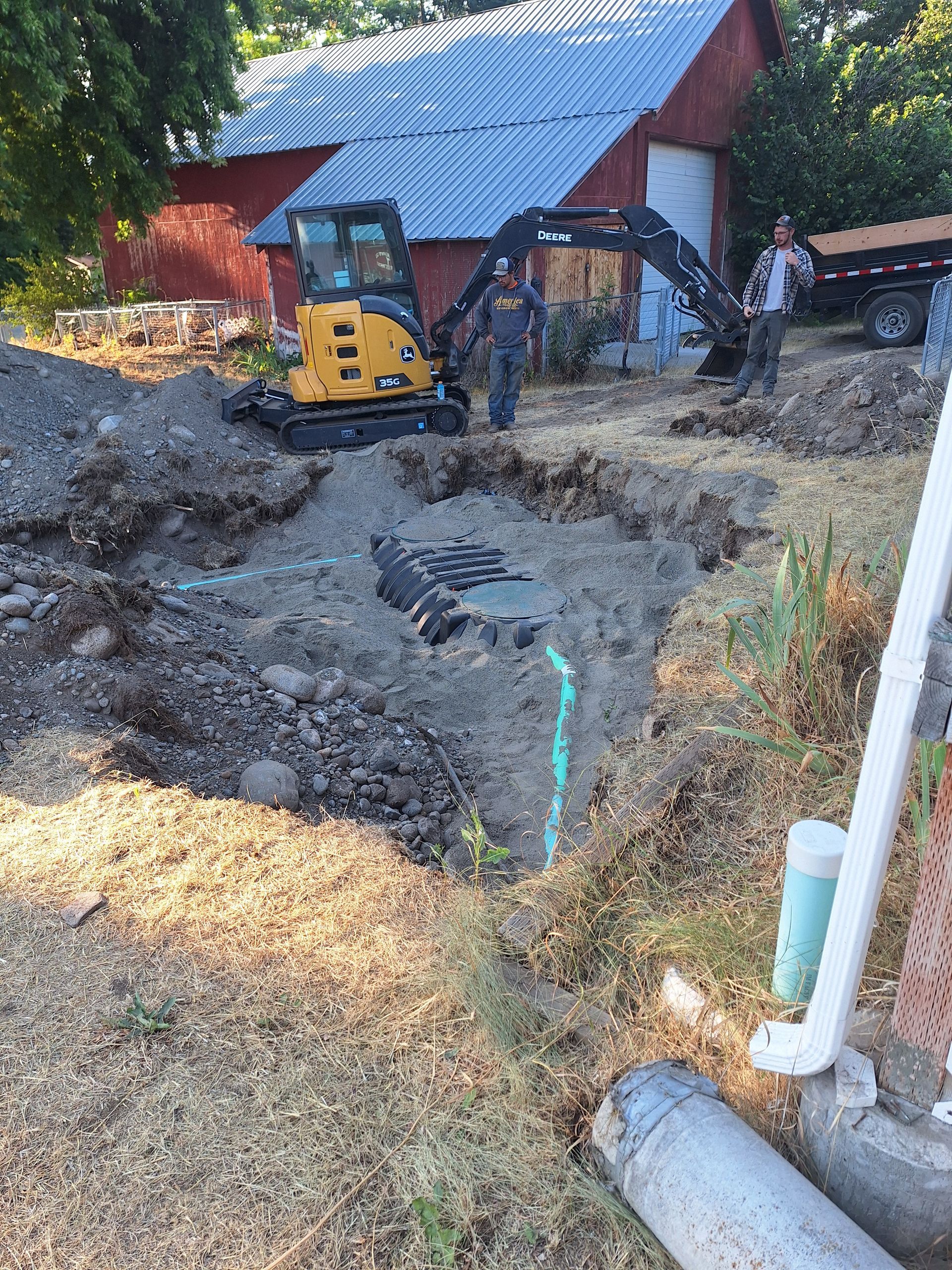 Excavator digging in front of a red barn; two people observe.