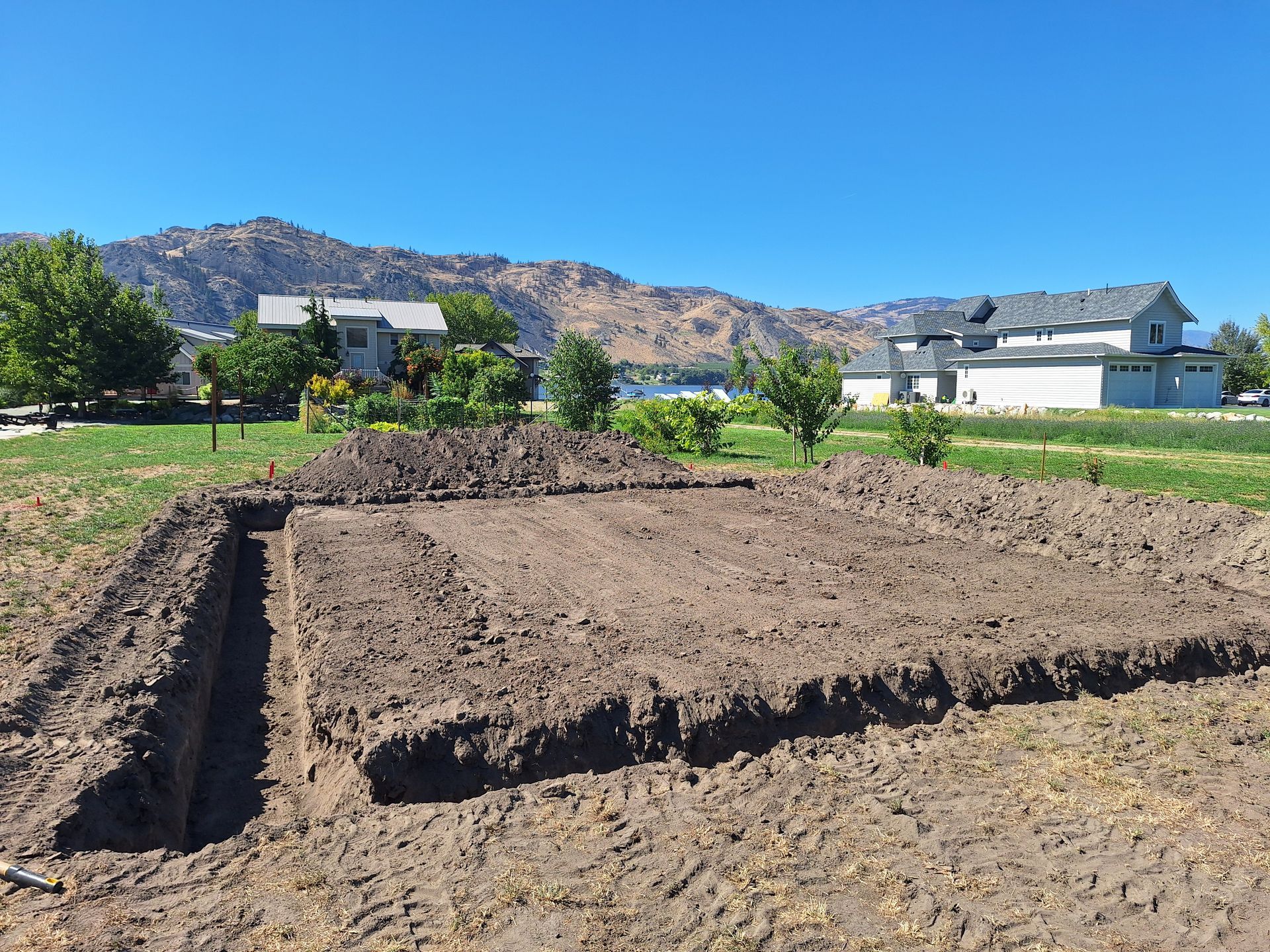 Excavated plot of land, brown dirt, bordered by a trench. Green grass, blue sky, houses, and mountains in background.