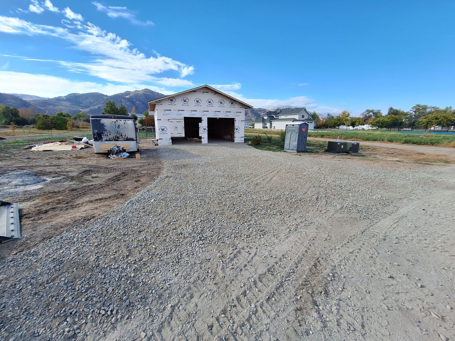 Garage under construction on a gravel driveway, with blue sky and mountains in the background.