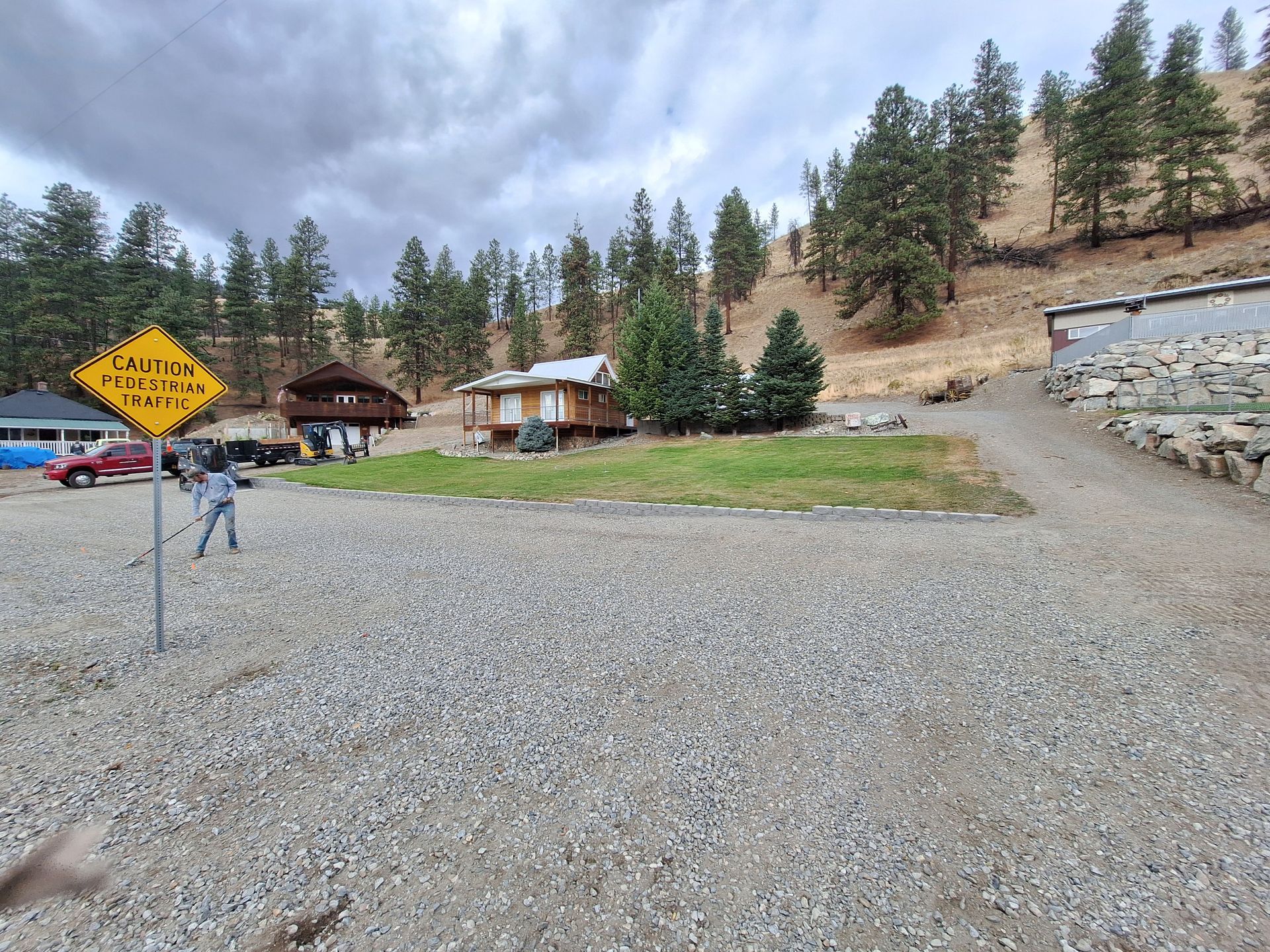 Gravel driveway leads to a wooden cabin and yard, with trees on a hillside under a cloudy sky.