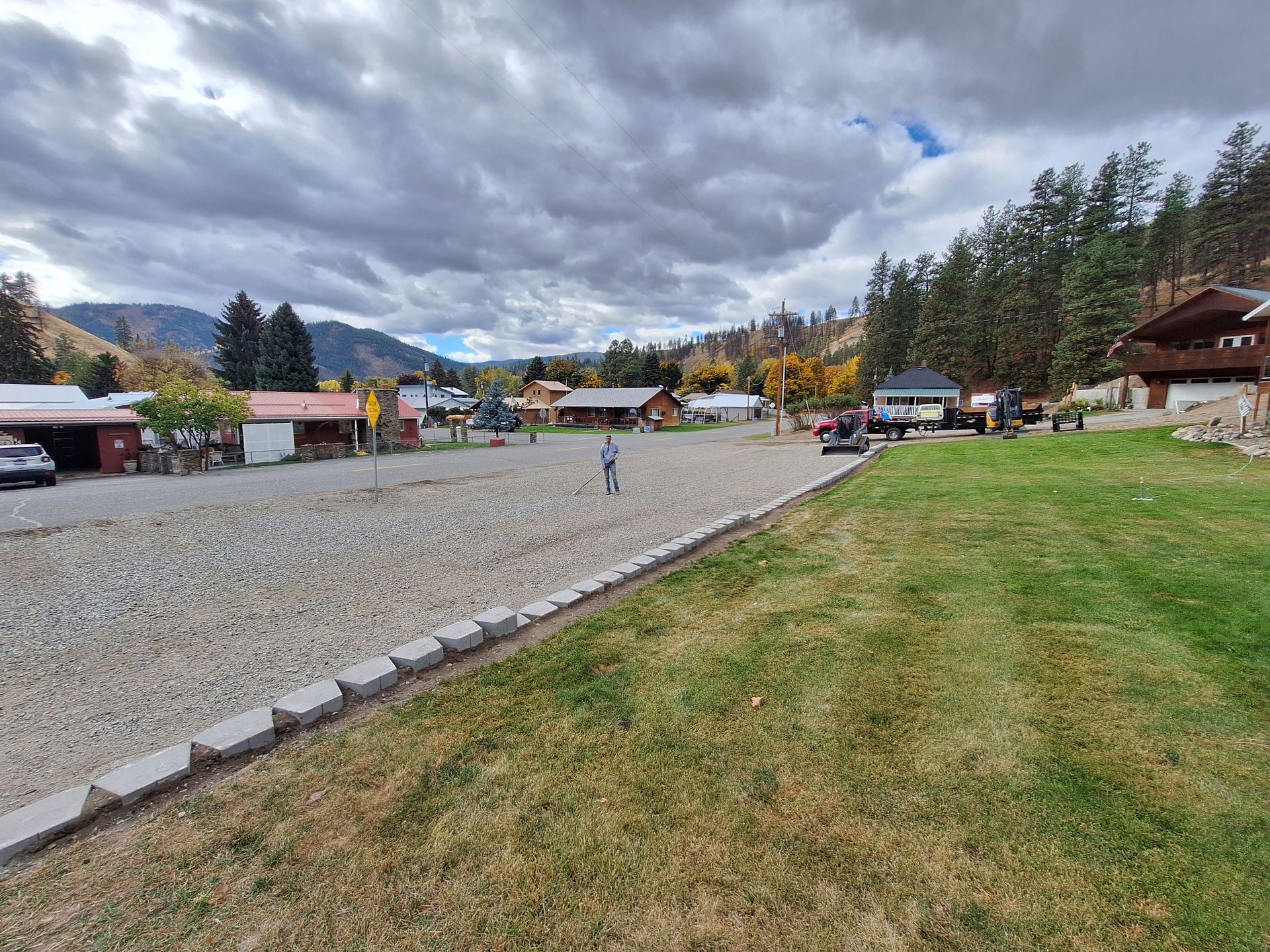 A gravel parking area with a green lawn. Buildings and trees in the background under a cloudy sky.