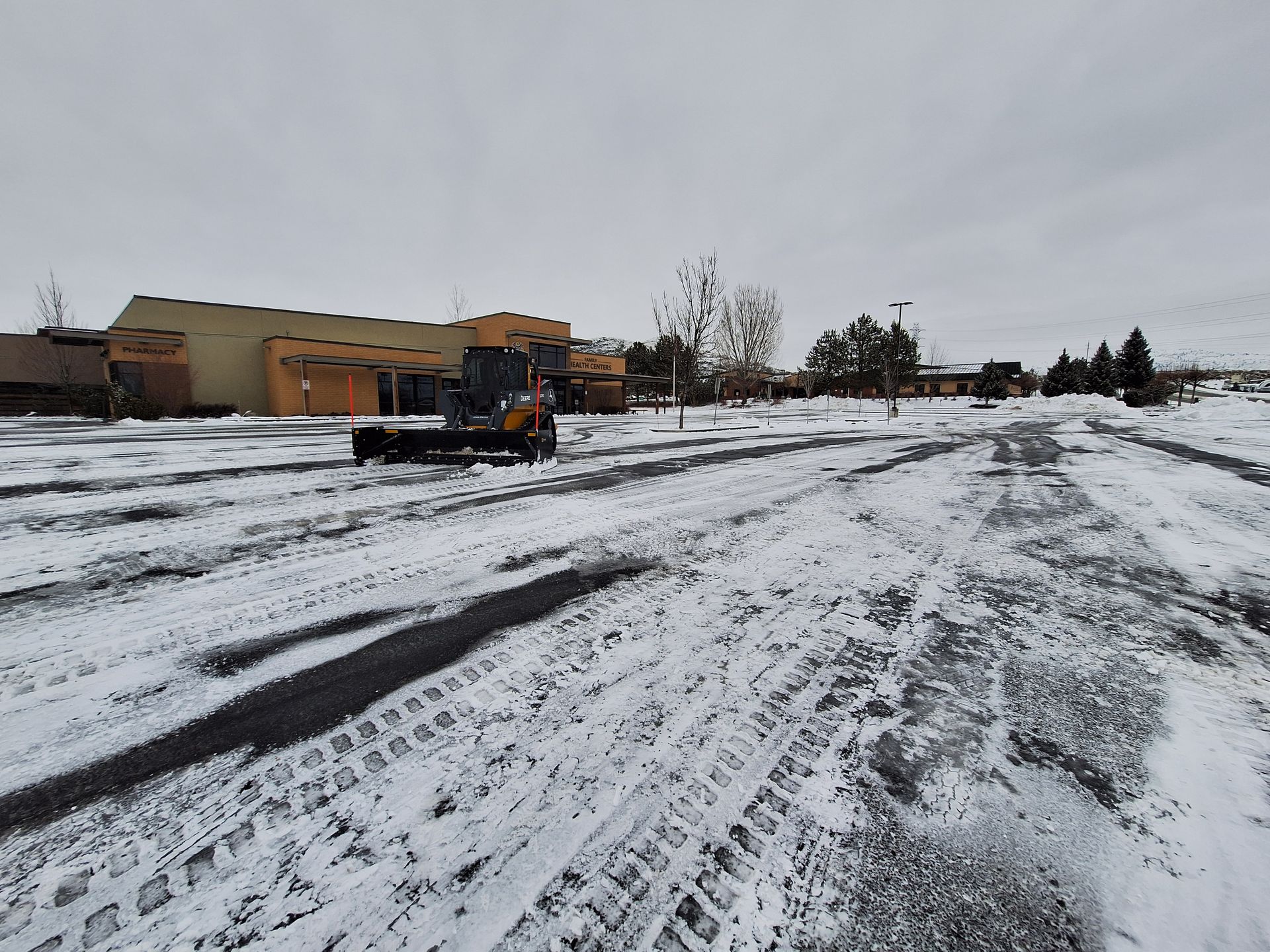 Snow-covered parking lot with a yellow loader clearing snow; low-angle shot, overcast sky, building in the background.