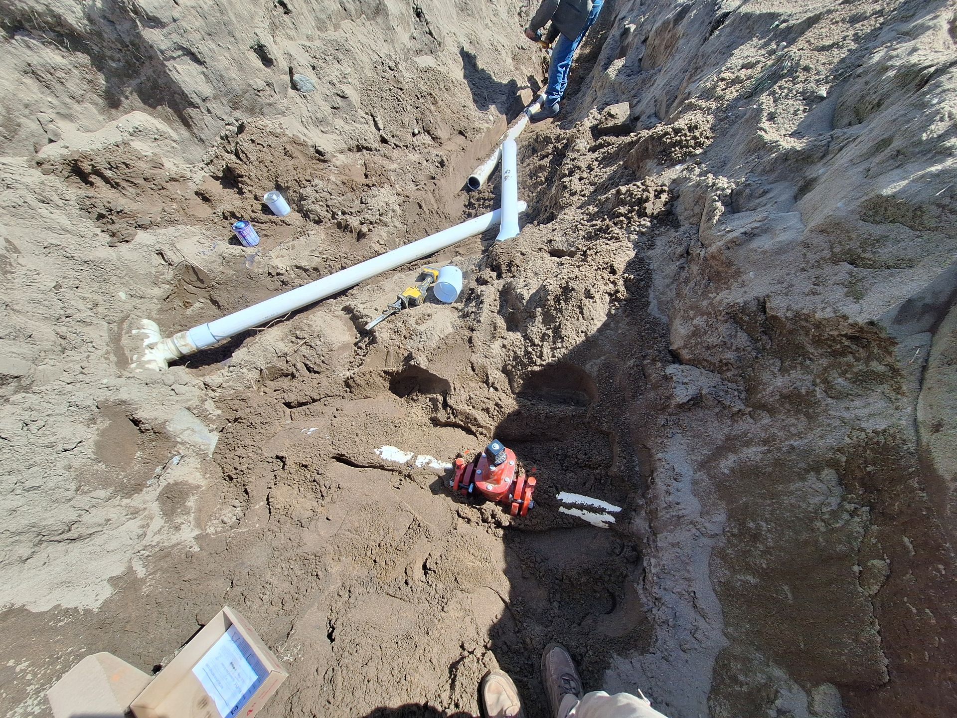 Trench with installed blue PVC pipes, connectors, and a red valve, in brown soil.