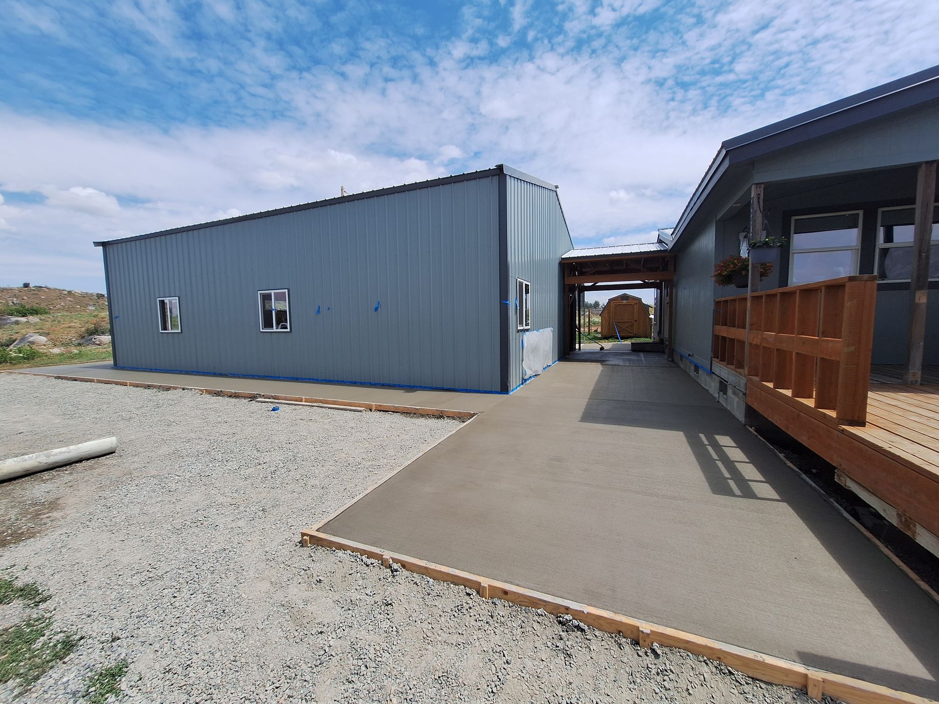 Exterior view of a blue metal building connected to a house with a concrete walkway and gravel ground.