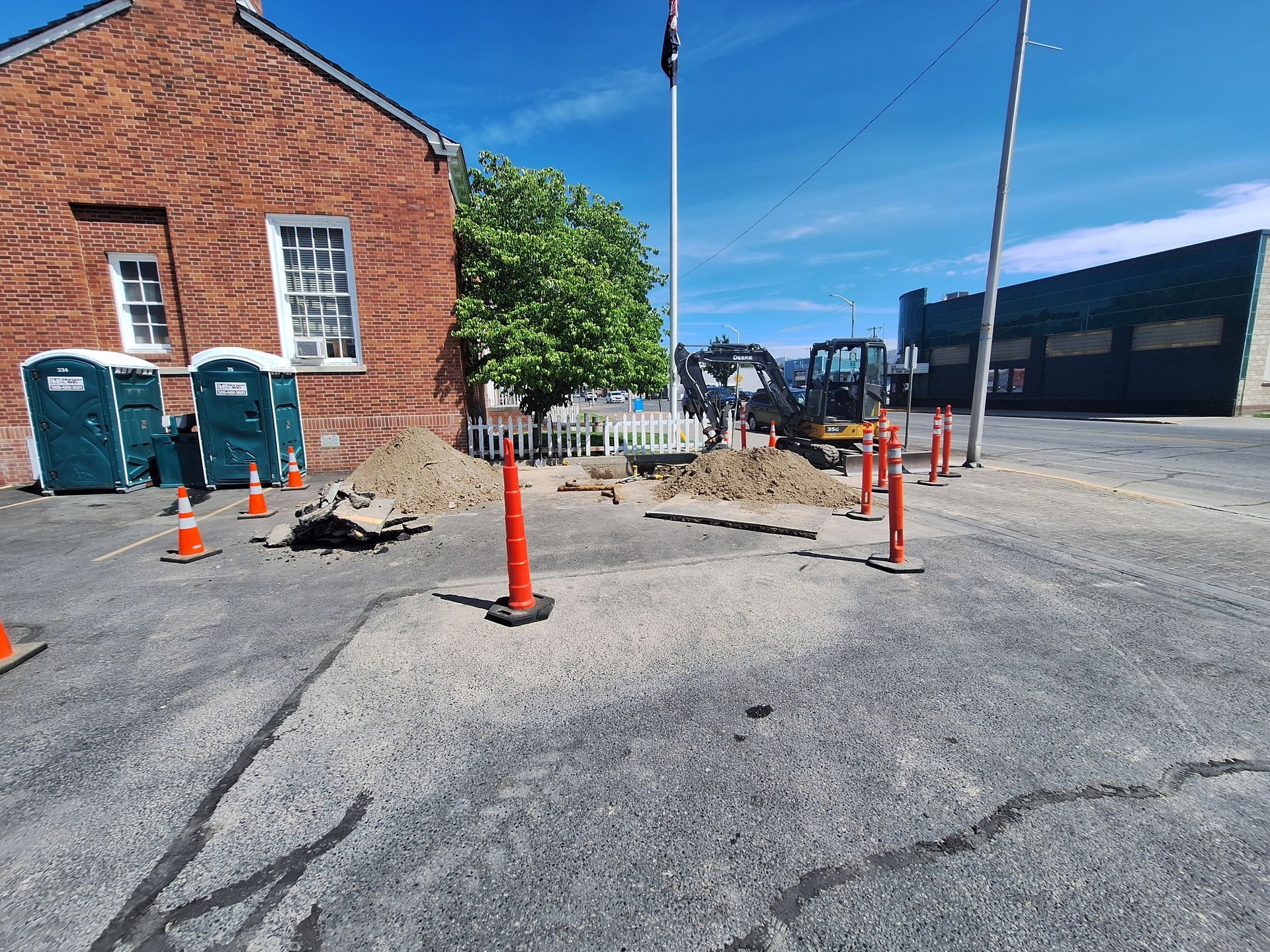 Construction site outside a brick building with a small excavator, piles of dirt, and portable toilets.