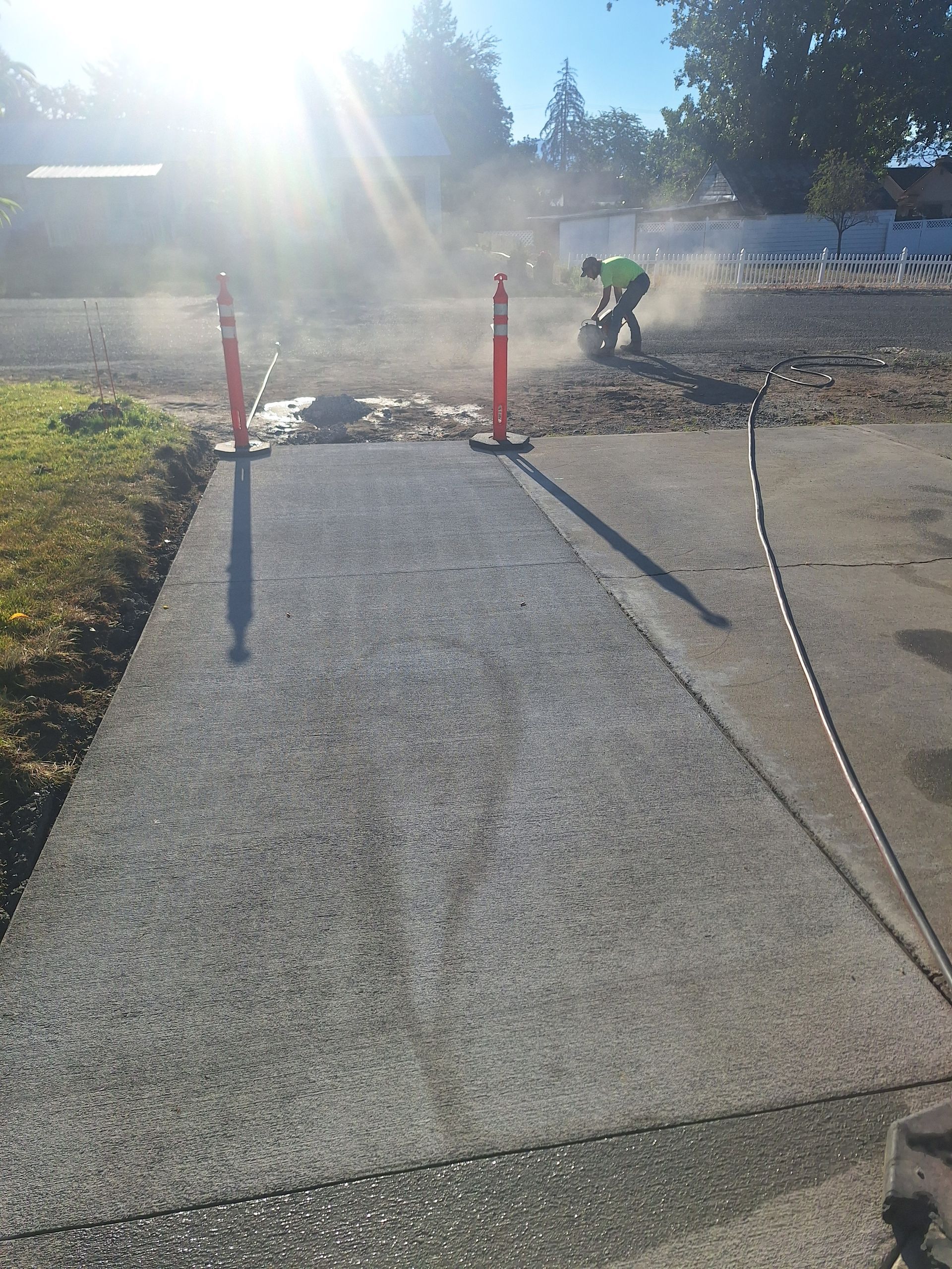 Asphalt paving in progress. Worker cuts existing pavement with saw, creating dust. Orange traffic cones mark the area.