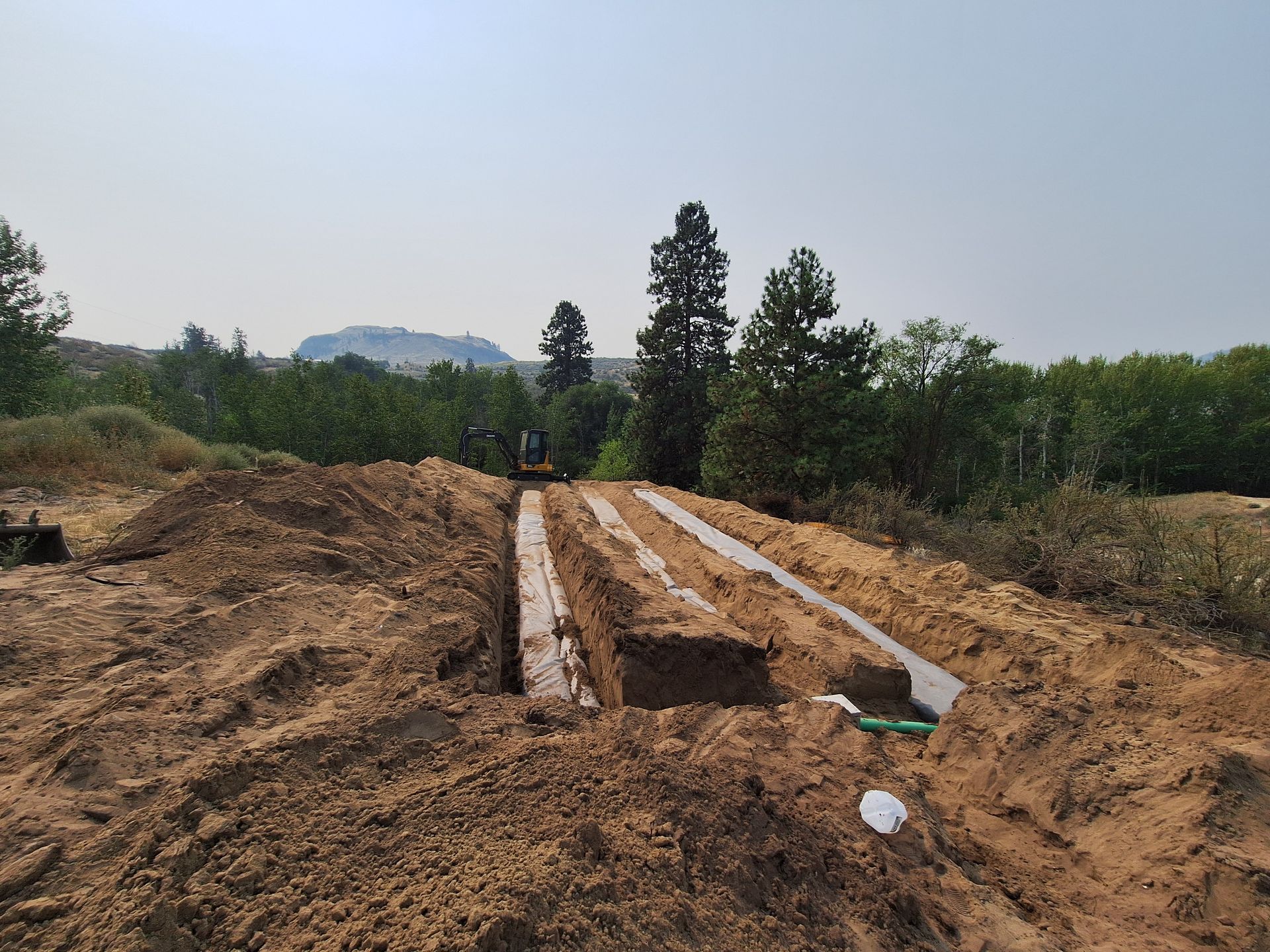 Excavated trenches for septic system in a dirt lot with trees and a hill in the background.