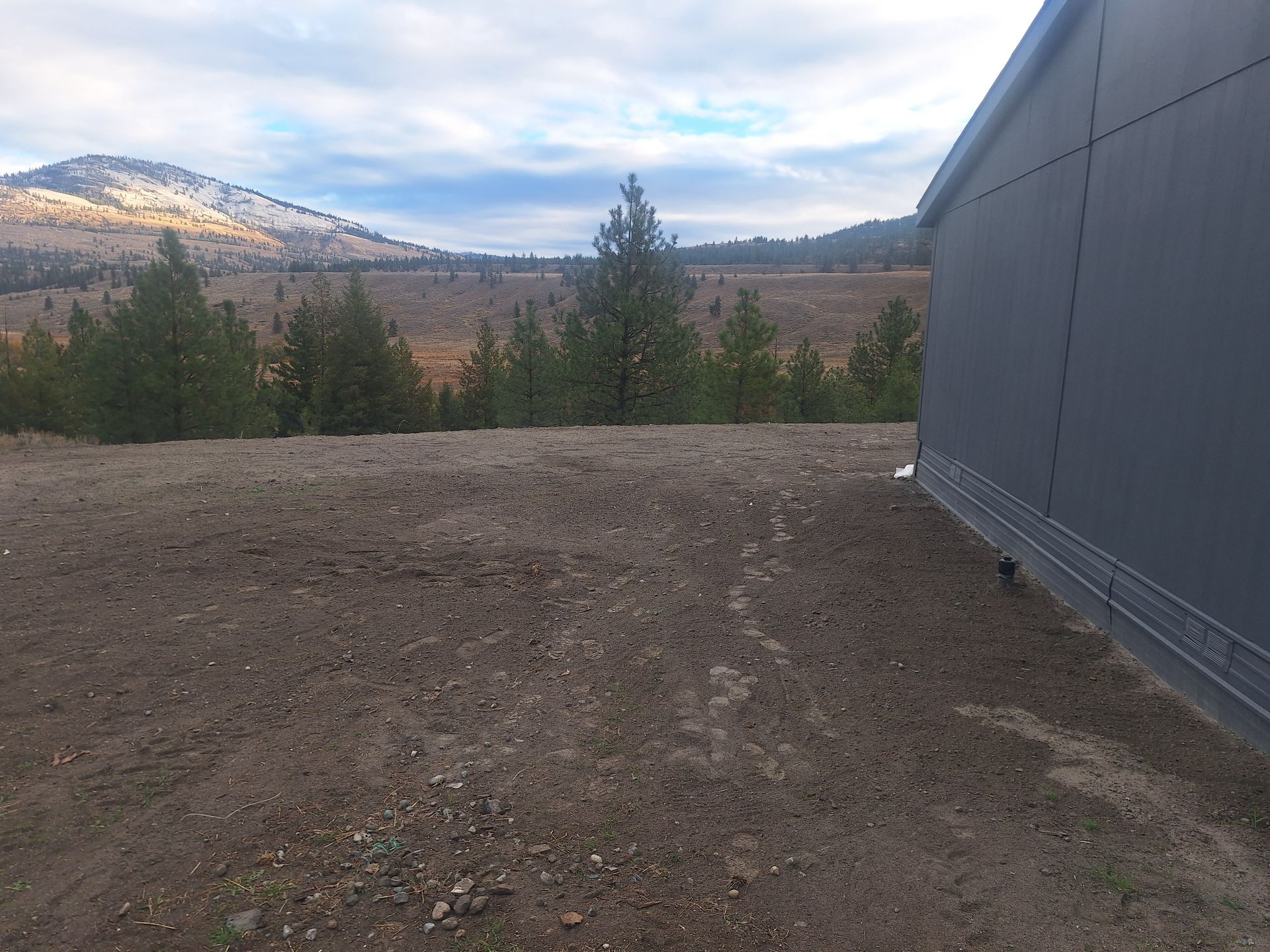 Dirt lot with a gray building on the right, trees, and a mountain range under a cloudy sky.