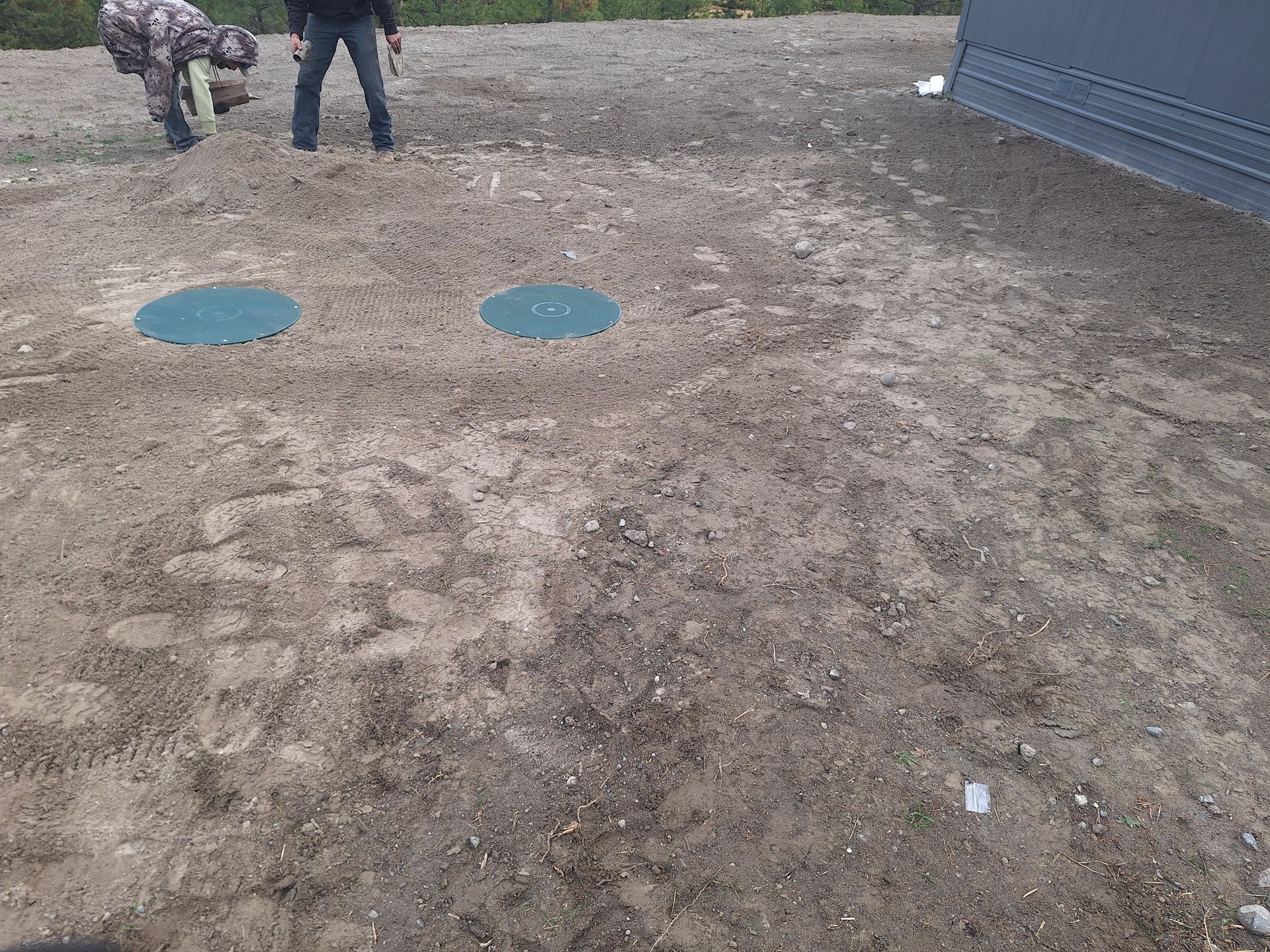 Two men near two green septic tank covers in a dirt lot next to a building.