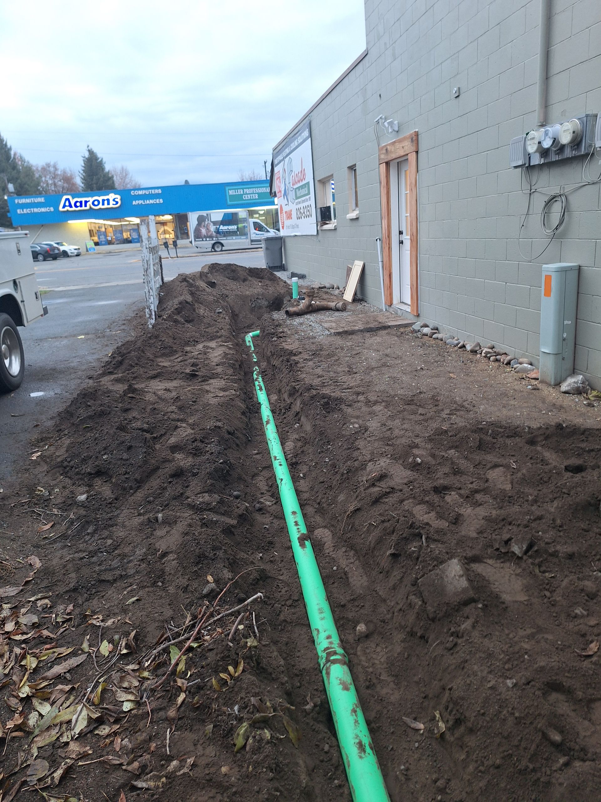 Trench dug alongside a building with green pipe running through it. Dirt, cars, and a blue sign are visible.