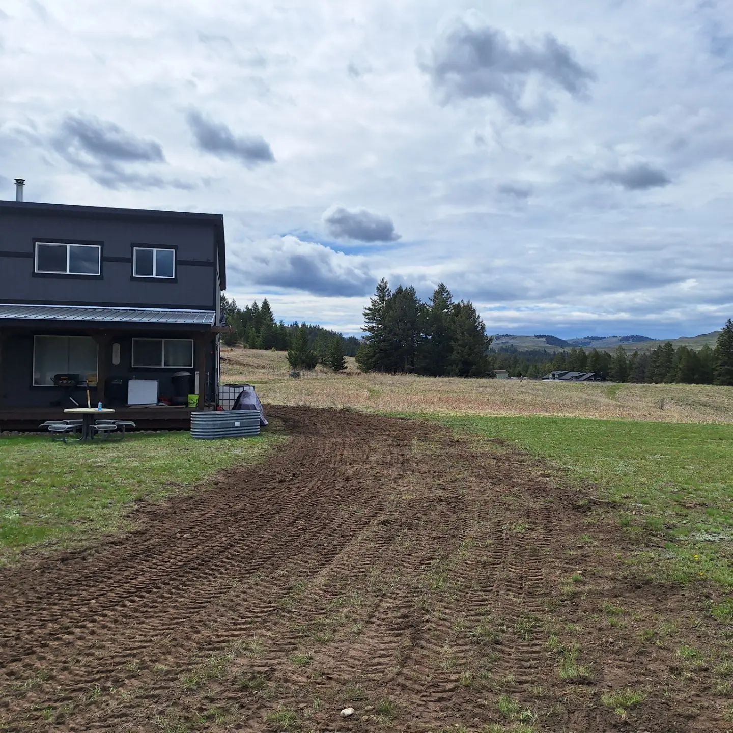 A dark-colored house with a yard. Tilled dirt leads into a field. Cloudy sky and distant trees.