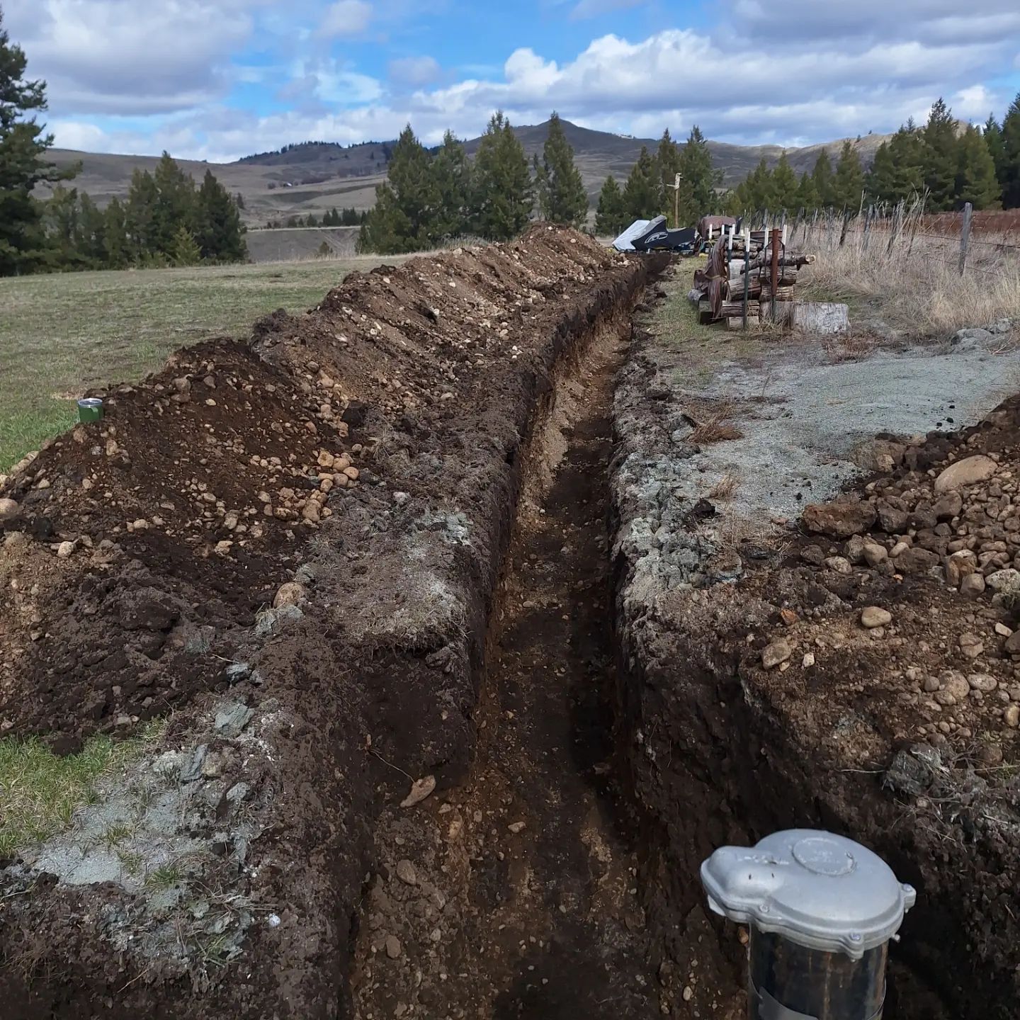 Long trench dug in field, leading to a wellhead. Dirt piles on either side, rolling hills in background.