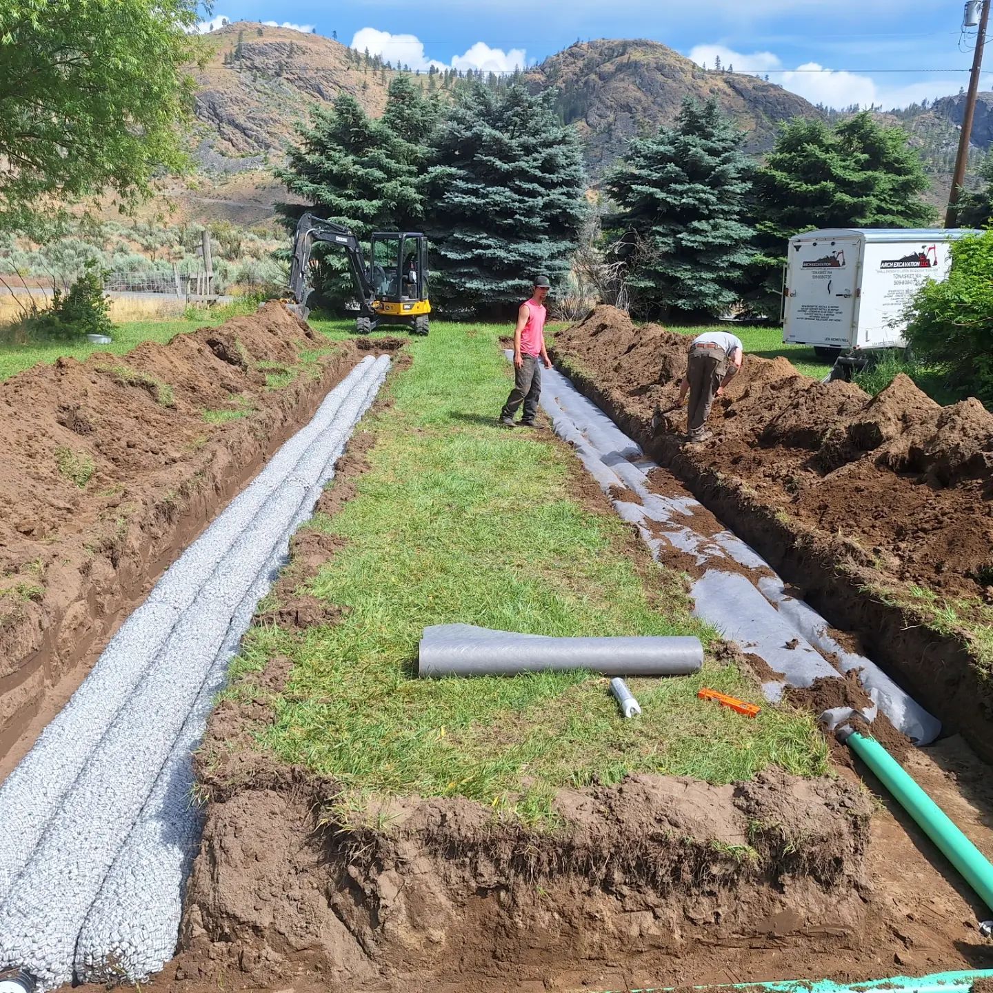 Construction site with trenches filled with gravel and fabric. Workers install drainage.