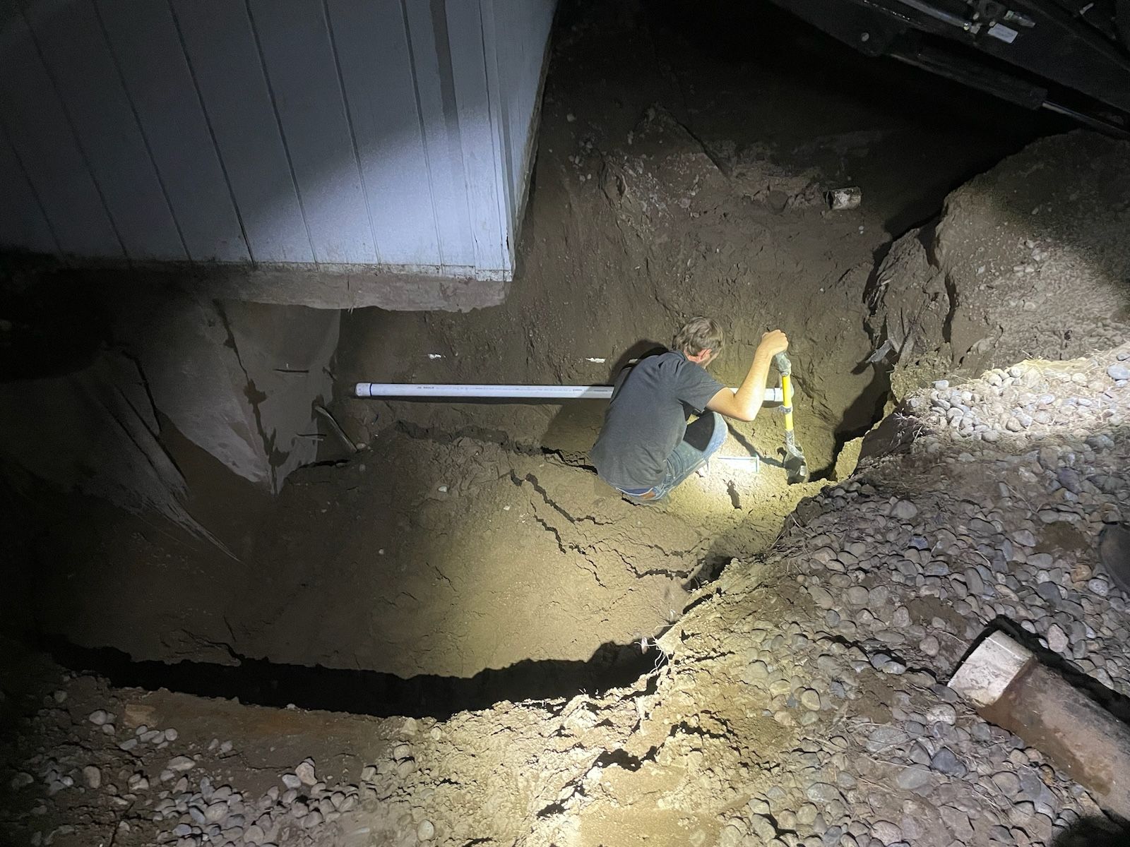Person in a trench near a building. Working with a pipe and measuring tools. Soil and gravel surround.