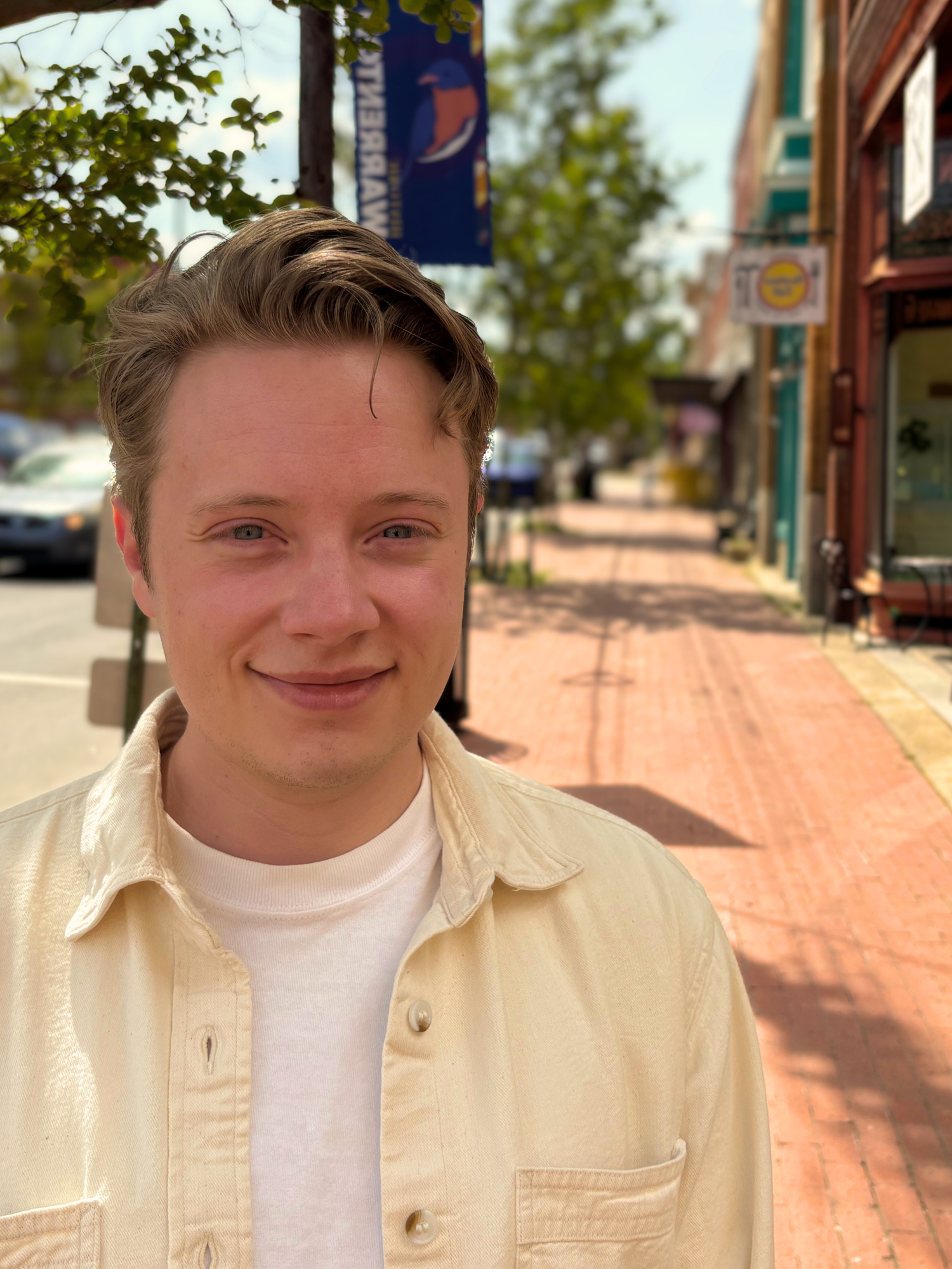 Person standing on a sunny brick sidewalk in front of shops, wearing a cream shirt and smiling slightly.