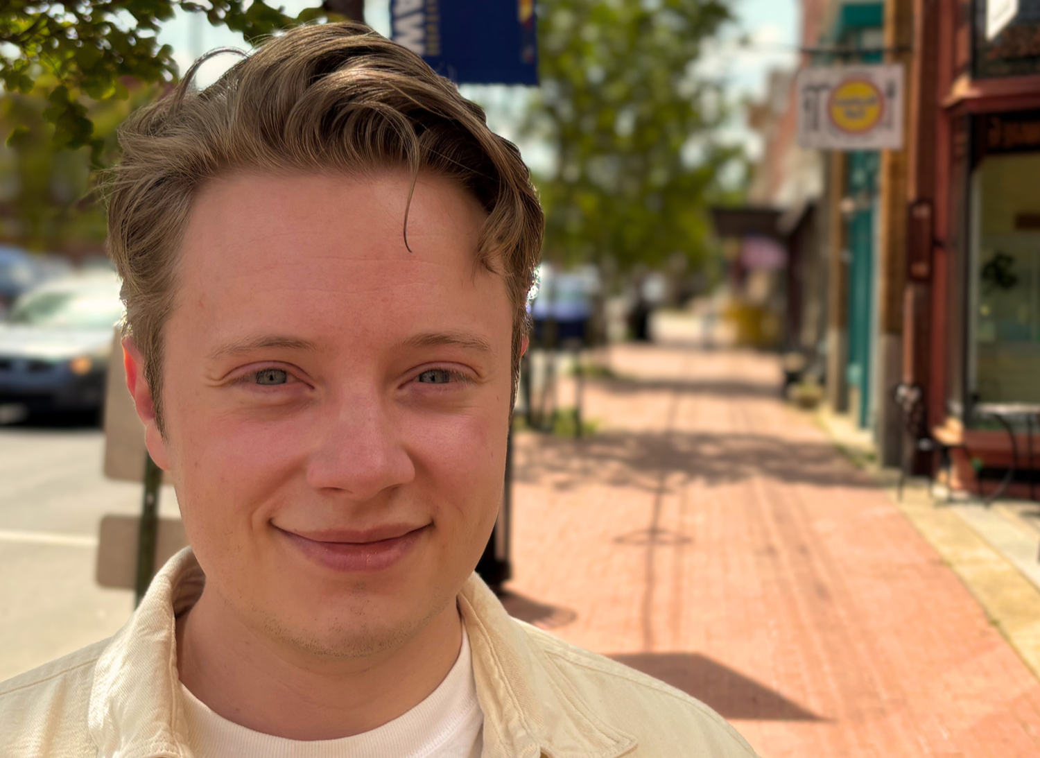 Smiling person on a sunny sidewalk with shops and trees in the background