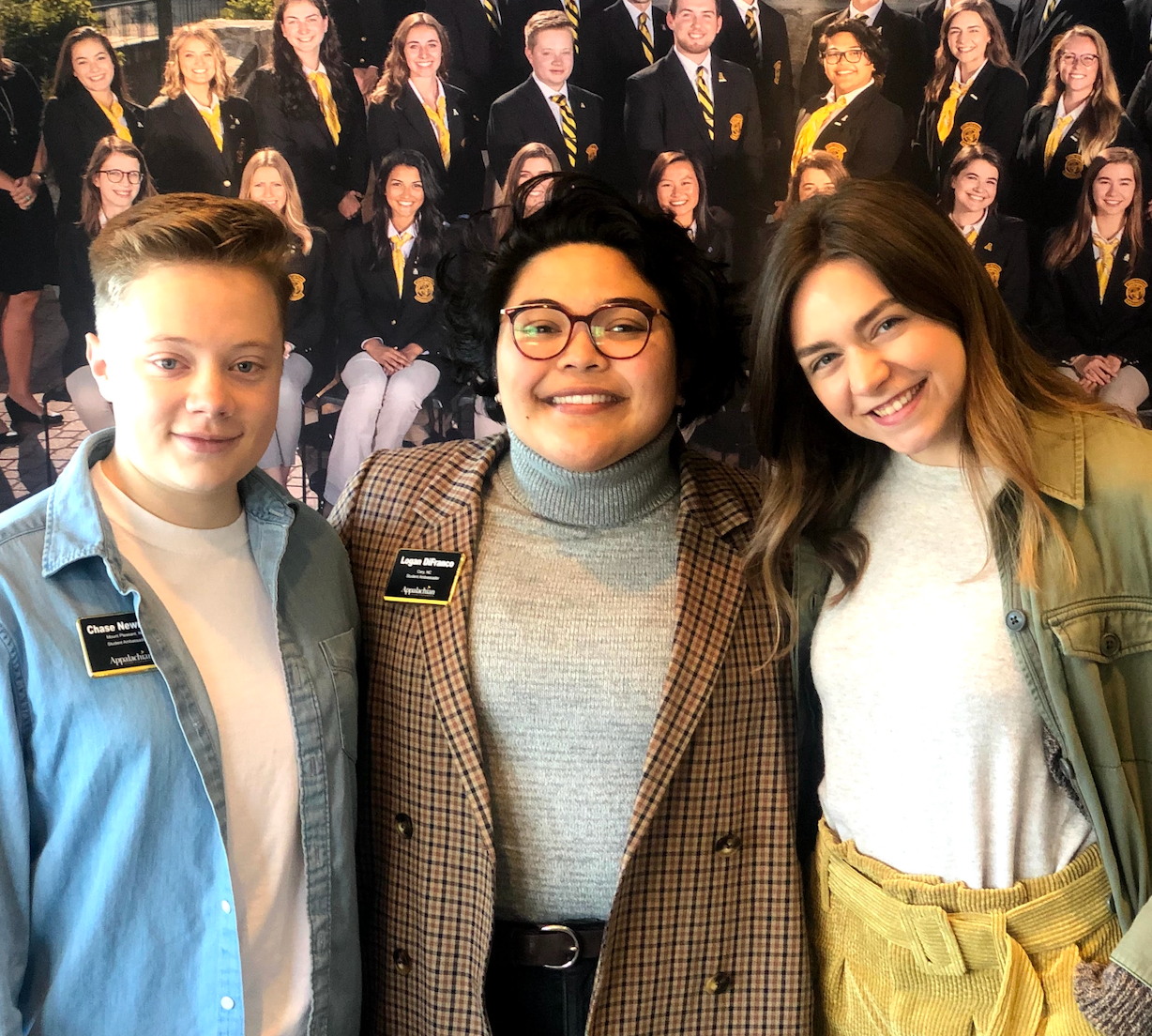 Three people smiling indoors, standing in front of a wall of graduation portraits.
