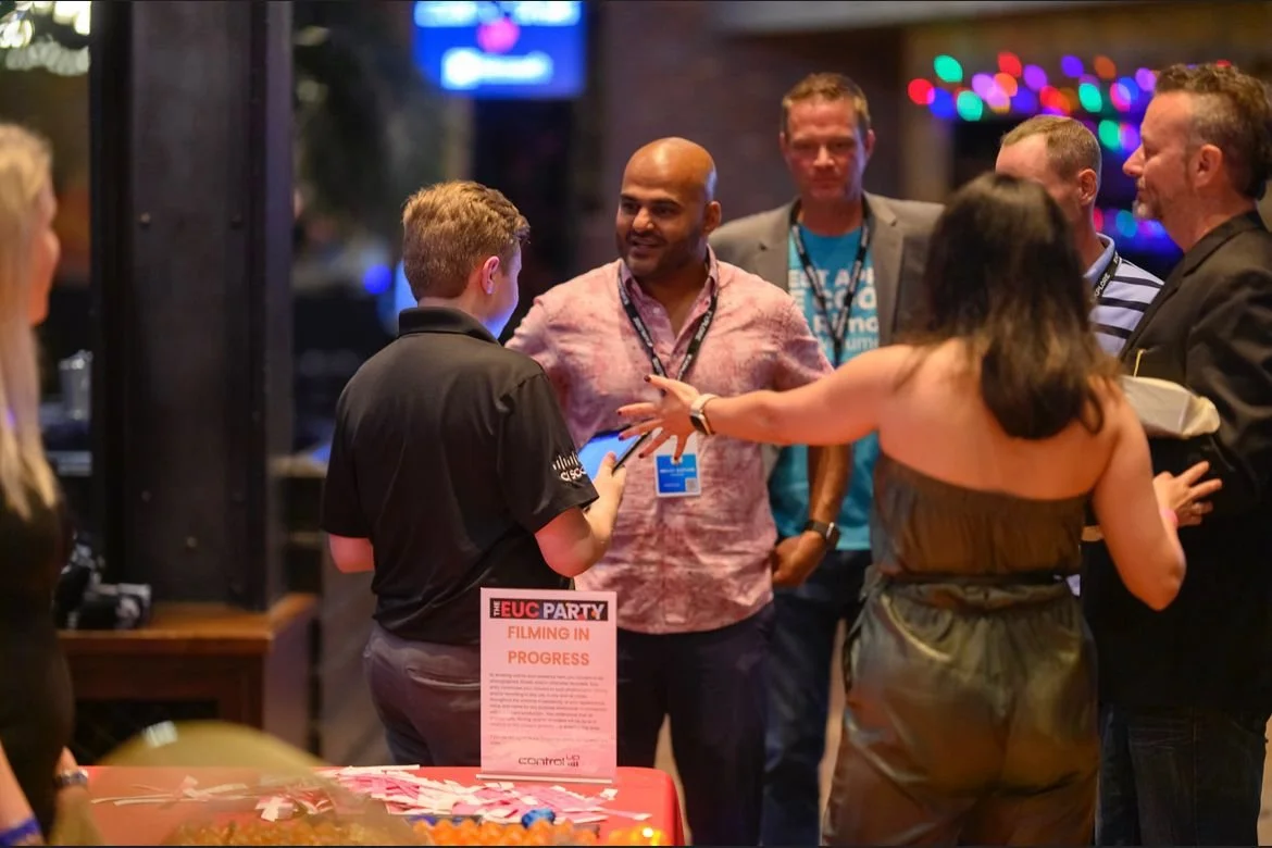 People mingling at a lively indoor event with colorful lights and a sign on a table