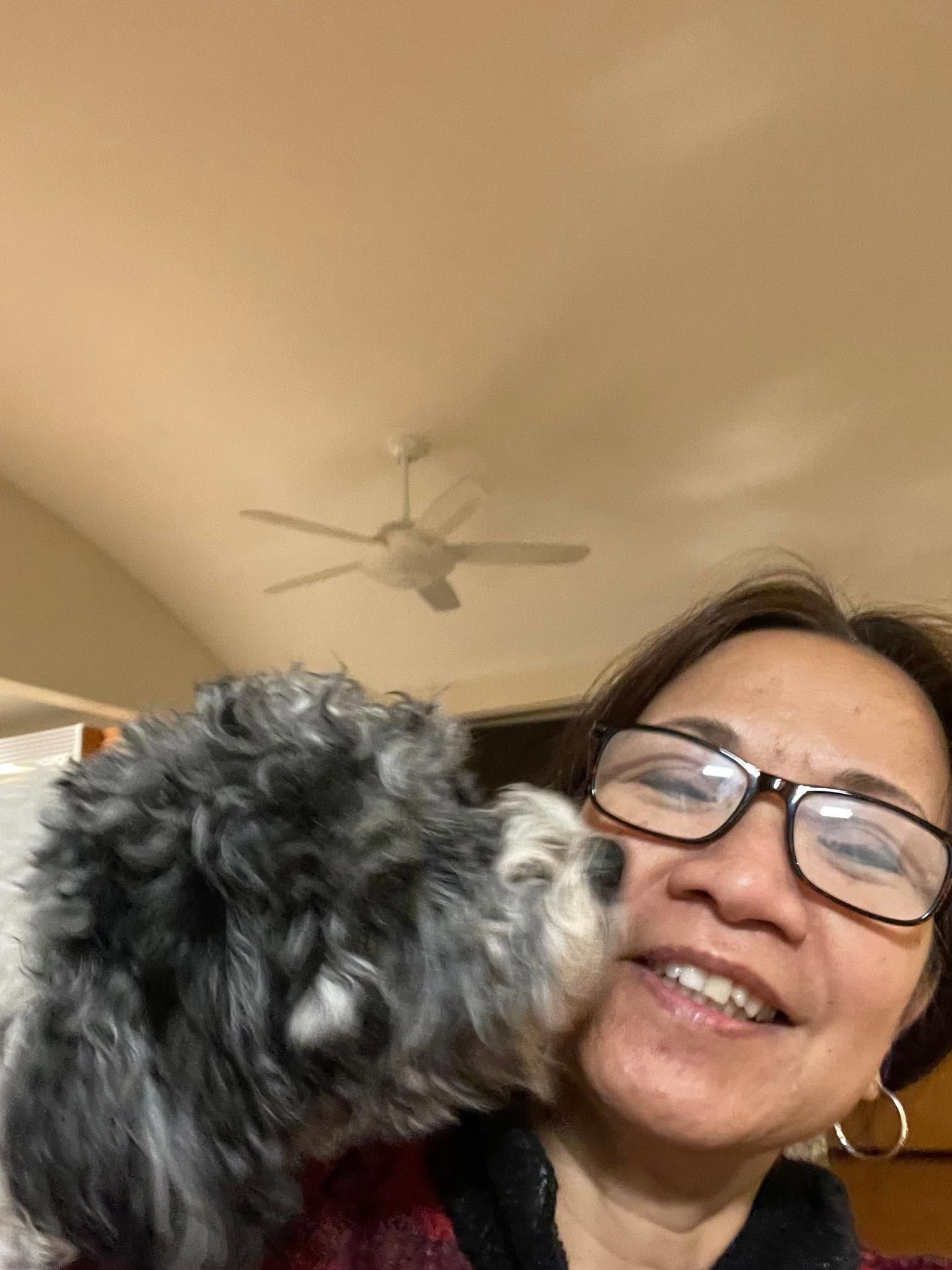 Woman smiling as a small black-and-gray dog nuzzles her cheek indoors.