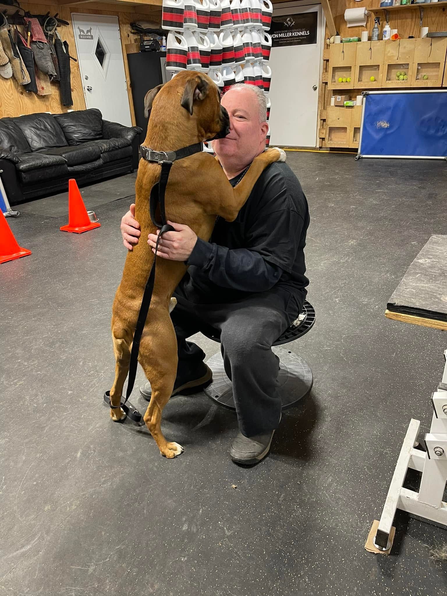 Man kneels and hugs a brown dog in a training room with cones and shelving.