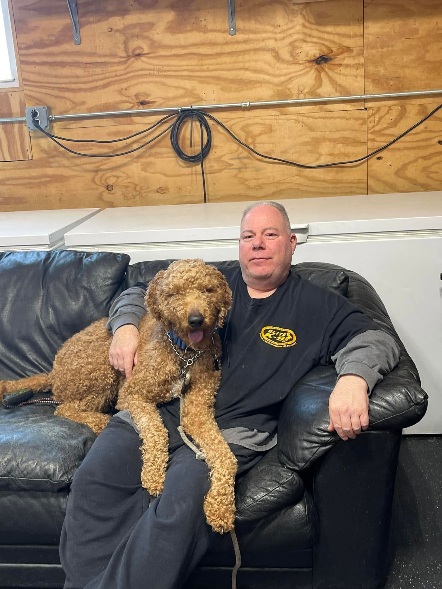 Man sitting on a black leather couch with a curly brown dog in a wood-paneled room.