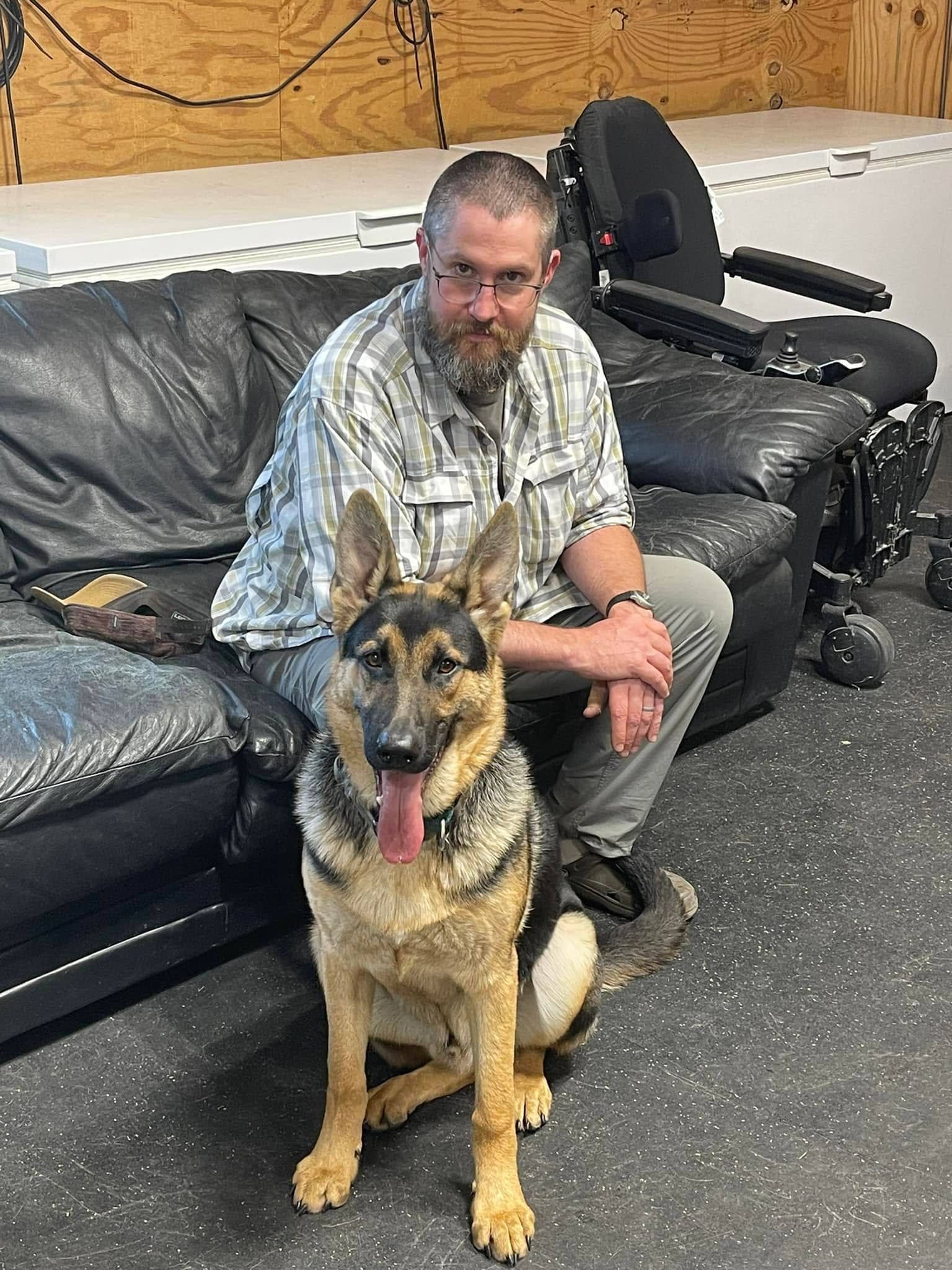 Man sitting on a black couch with a German shepherd in a room with carpet and office equipment.