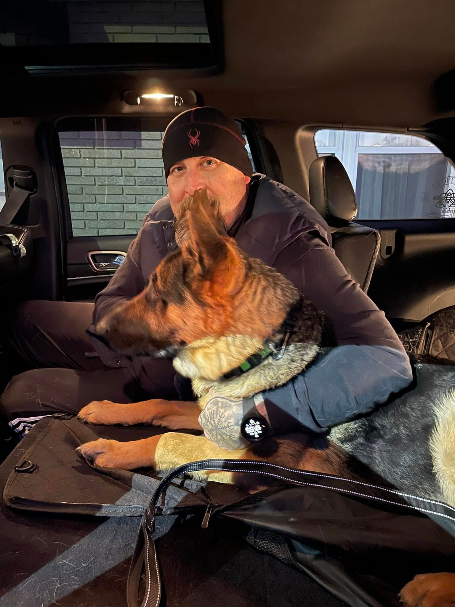 Man sitting in a car holding a German shepherd, both wearing winter gear