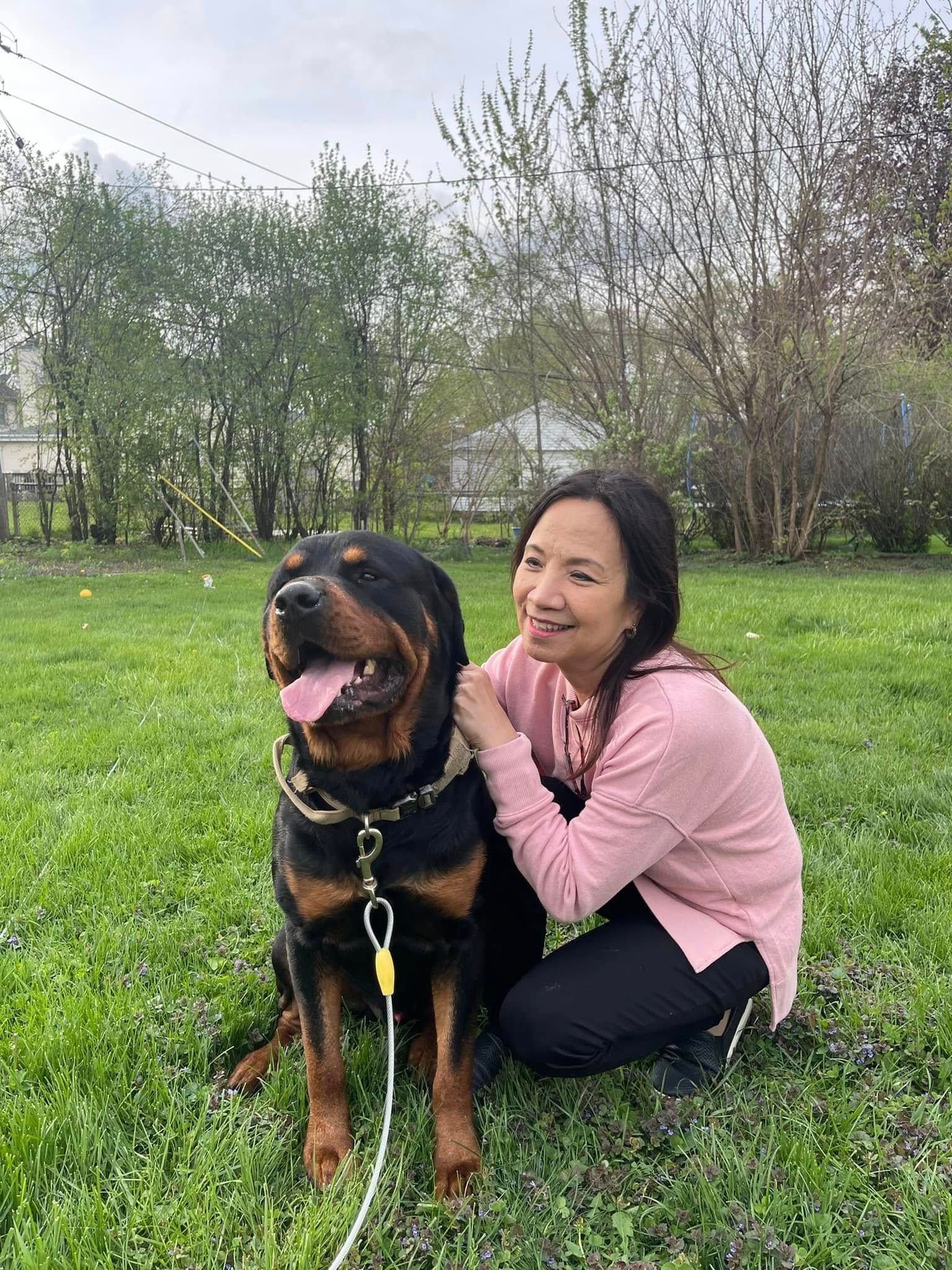 Woman crouching beside a large black and tan dog in a grassy park, both looking happy.
