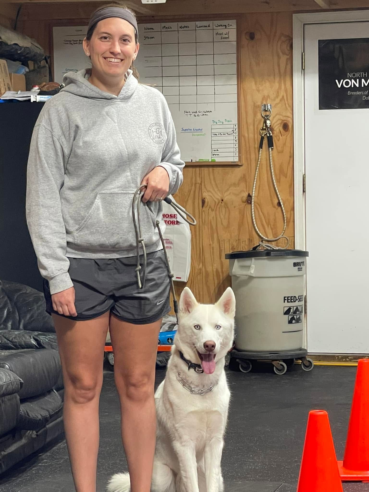 Woman standing indoors with a white dog beside her, both facing the camera in a gym or training space.