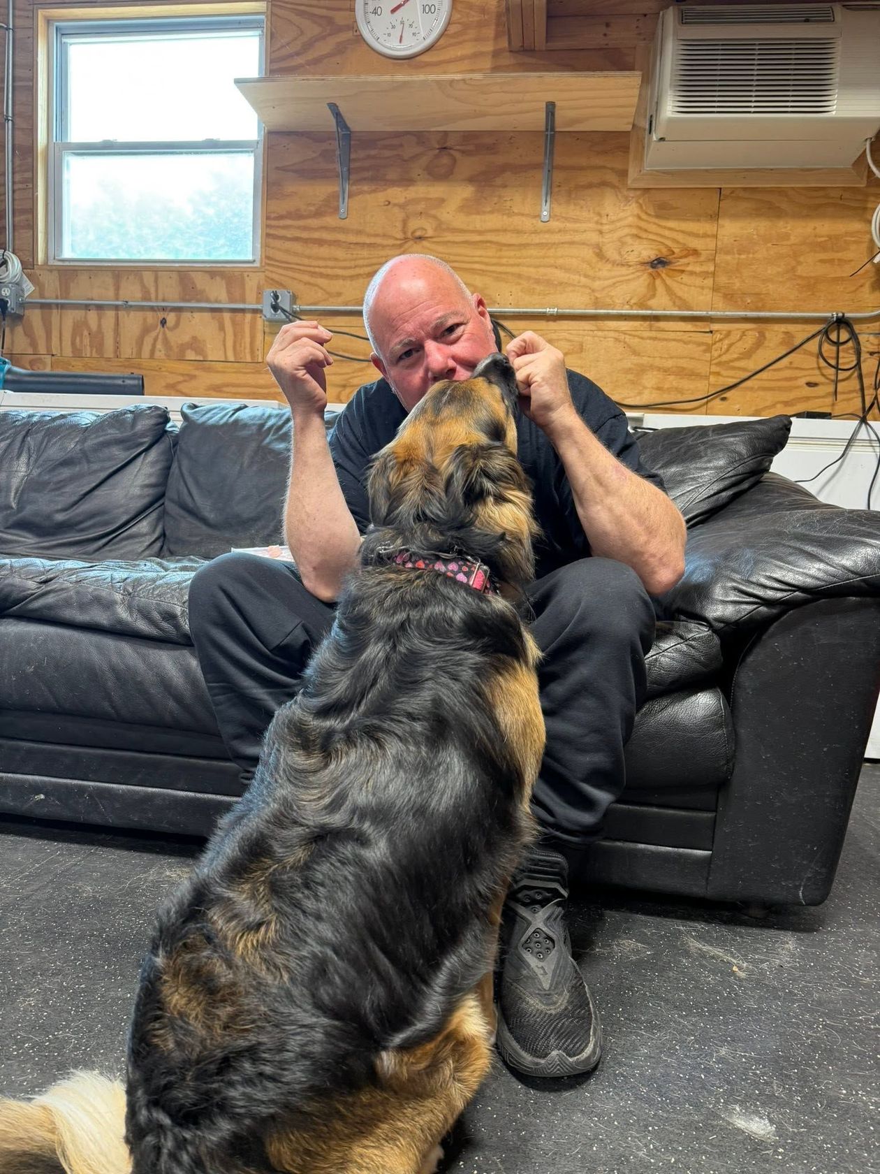 Man sitting on a couch while a large dog stands on its hind legs to nuzzle his face in a garage-like room
