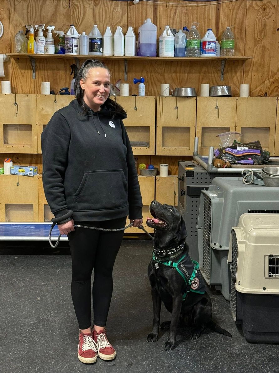 Woman standing beside a black dog in a training room with shelves and crates in the background