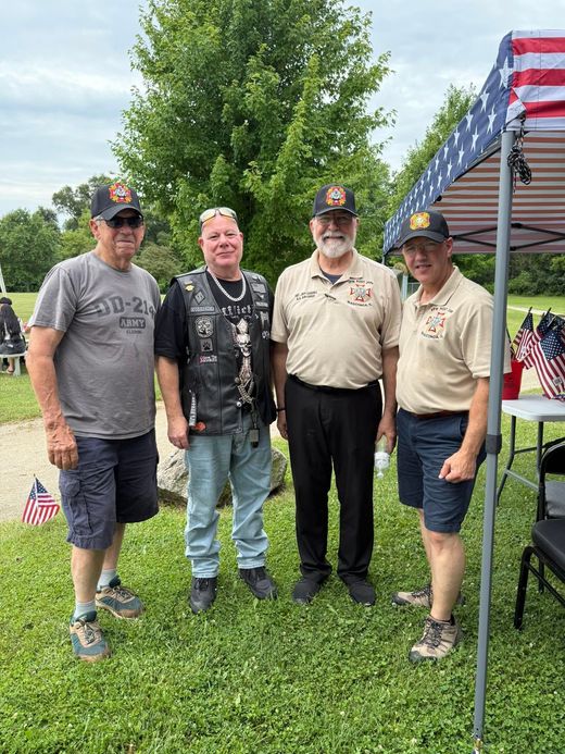 Four people standing on grass at an outdoor gathering beside a tent and American flags.