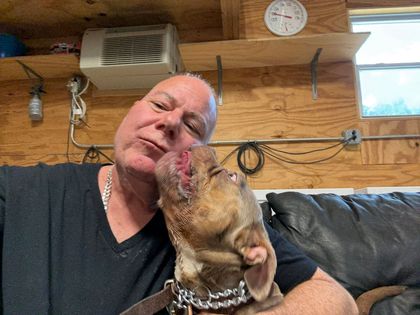 Man kissing a tan dog on the muzzle in a wooden room with a black couch and clock on the wall