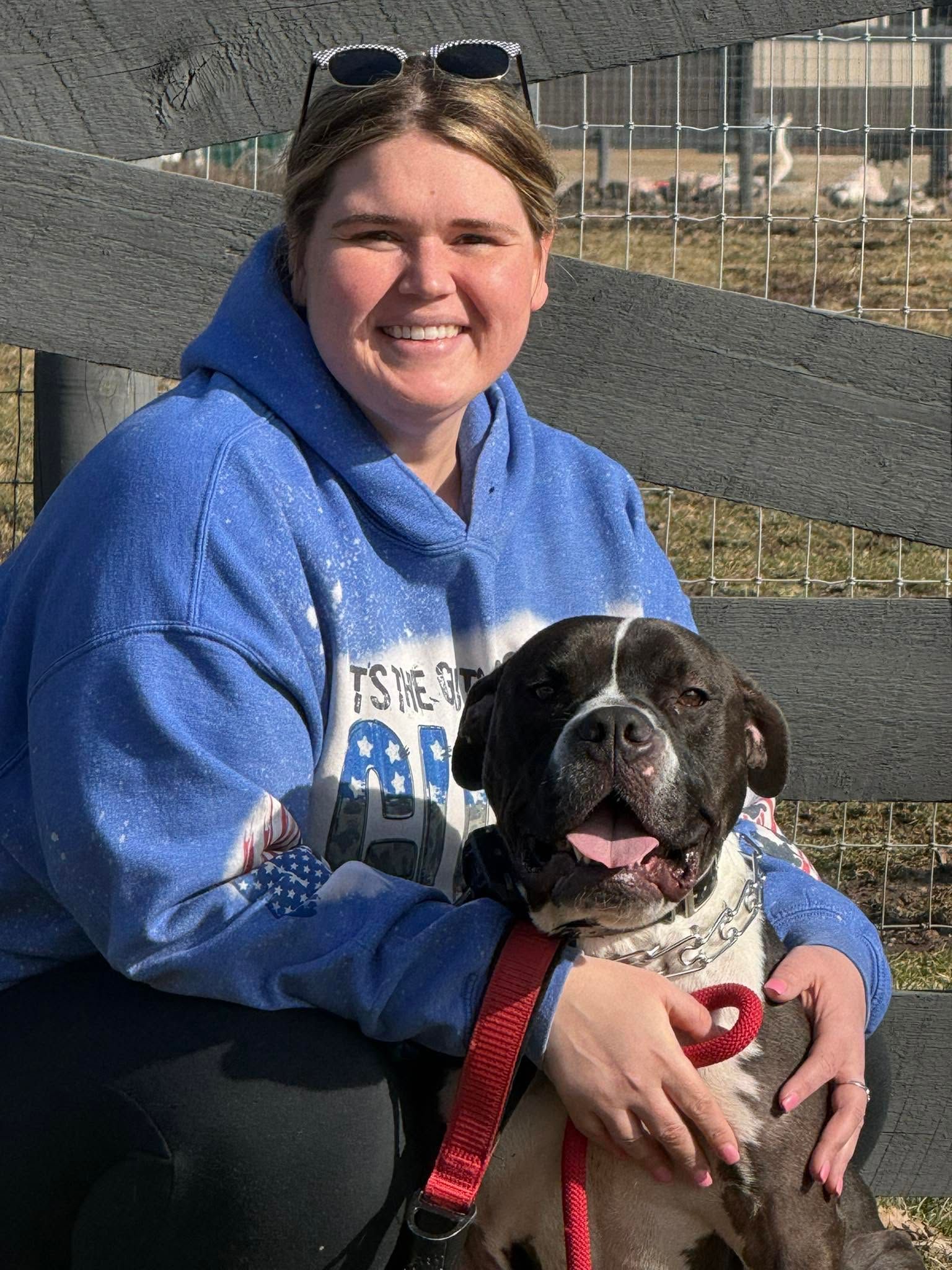 Smiling woman in a blue hoodie sitting with a black-and-white dog on a red leash outdoors