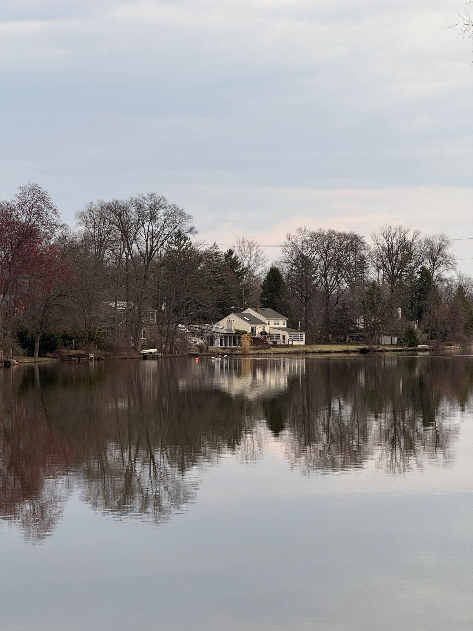 Calm lake reflecting bare trees and a small white house on the opposite shore