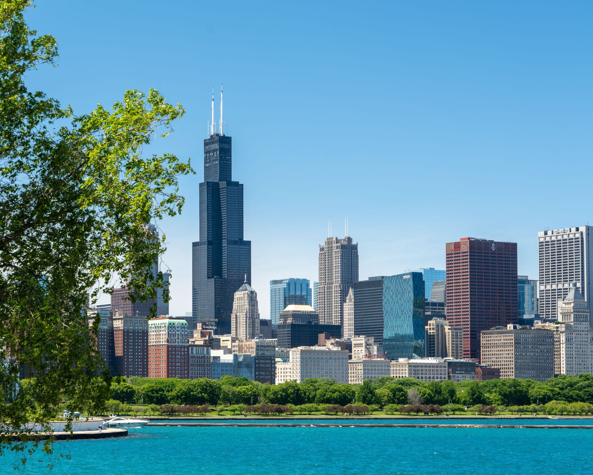 Chicago skyline with the Willis Tower behind turquoise water and green trees under a clear blue sky