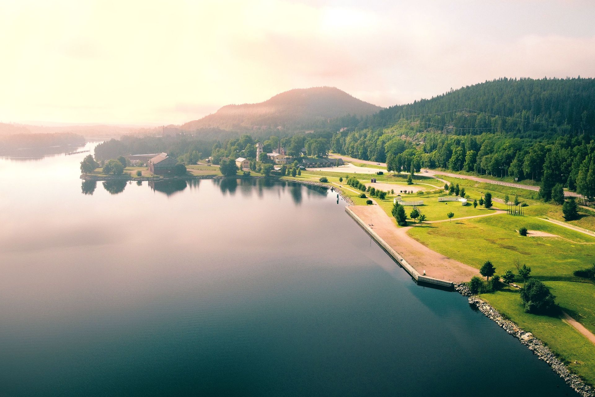 Aerial view of a calm lake beside a green park, forest, and hills at sunrise