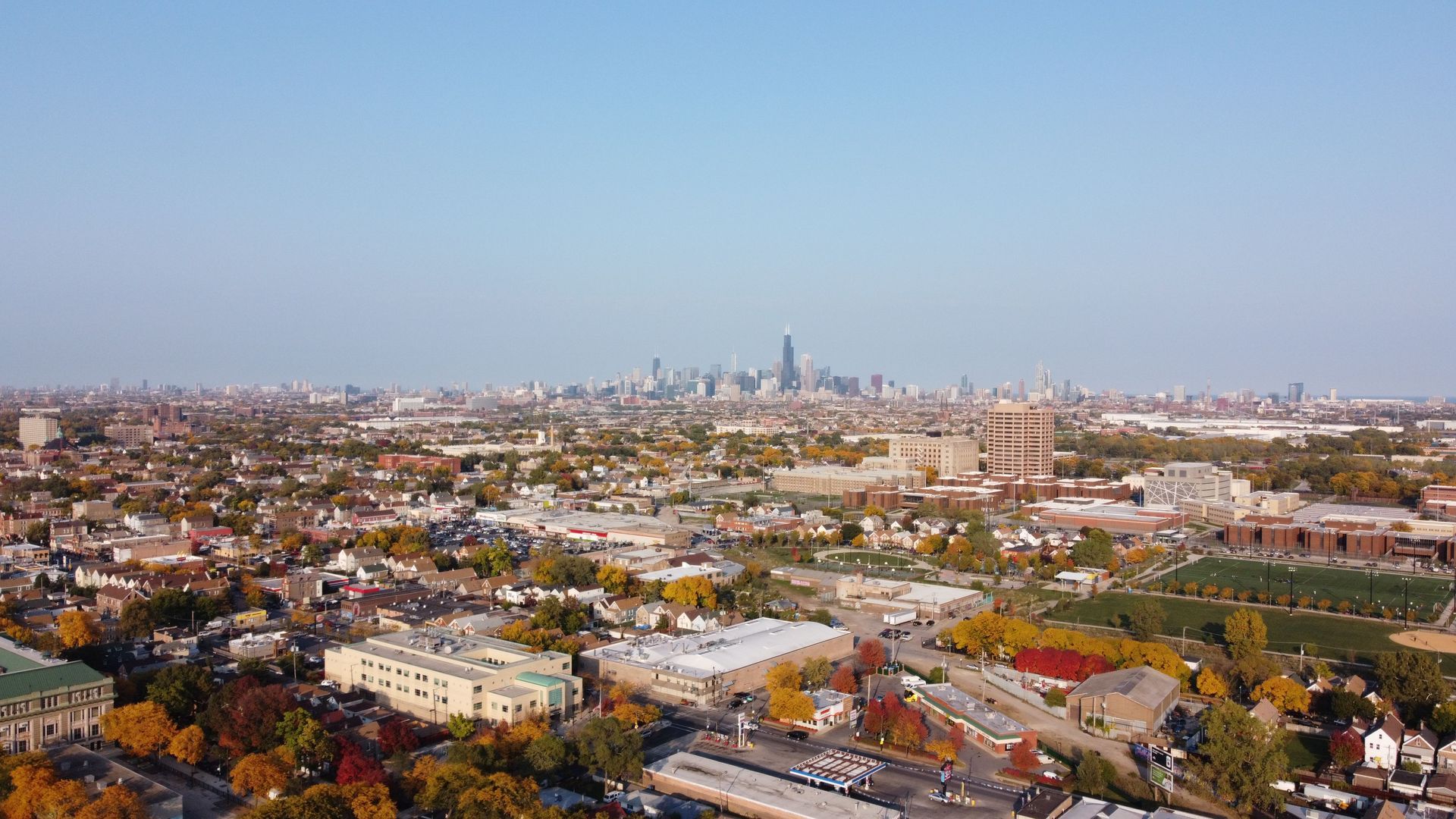 Aerial view of a city skyline with low-rise buildings and a distant cluster of skyscrapers under a clear blue sky