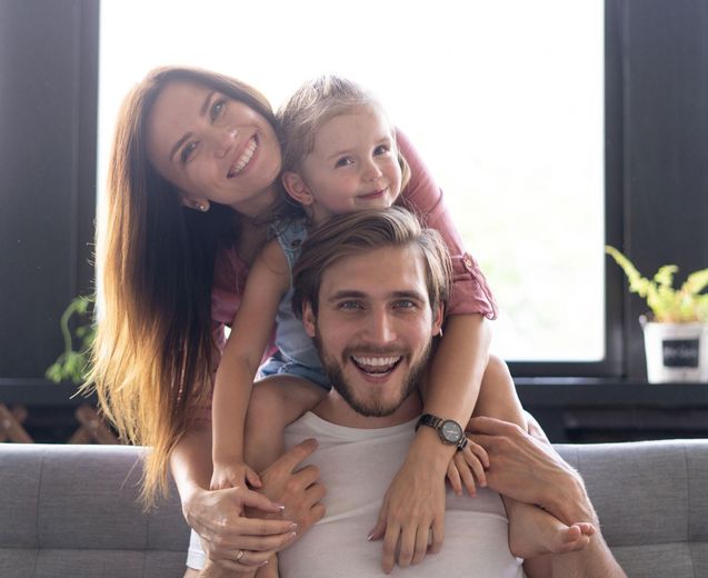 Smiling family on a couch, with a child on a man's shoulders and a woman leaning in behind them.