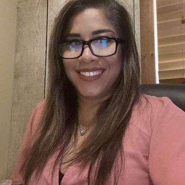 Smiling woman in glasses and a pink top, seated indoors against a wood-paneled wall.