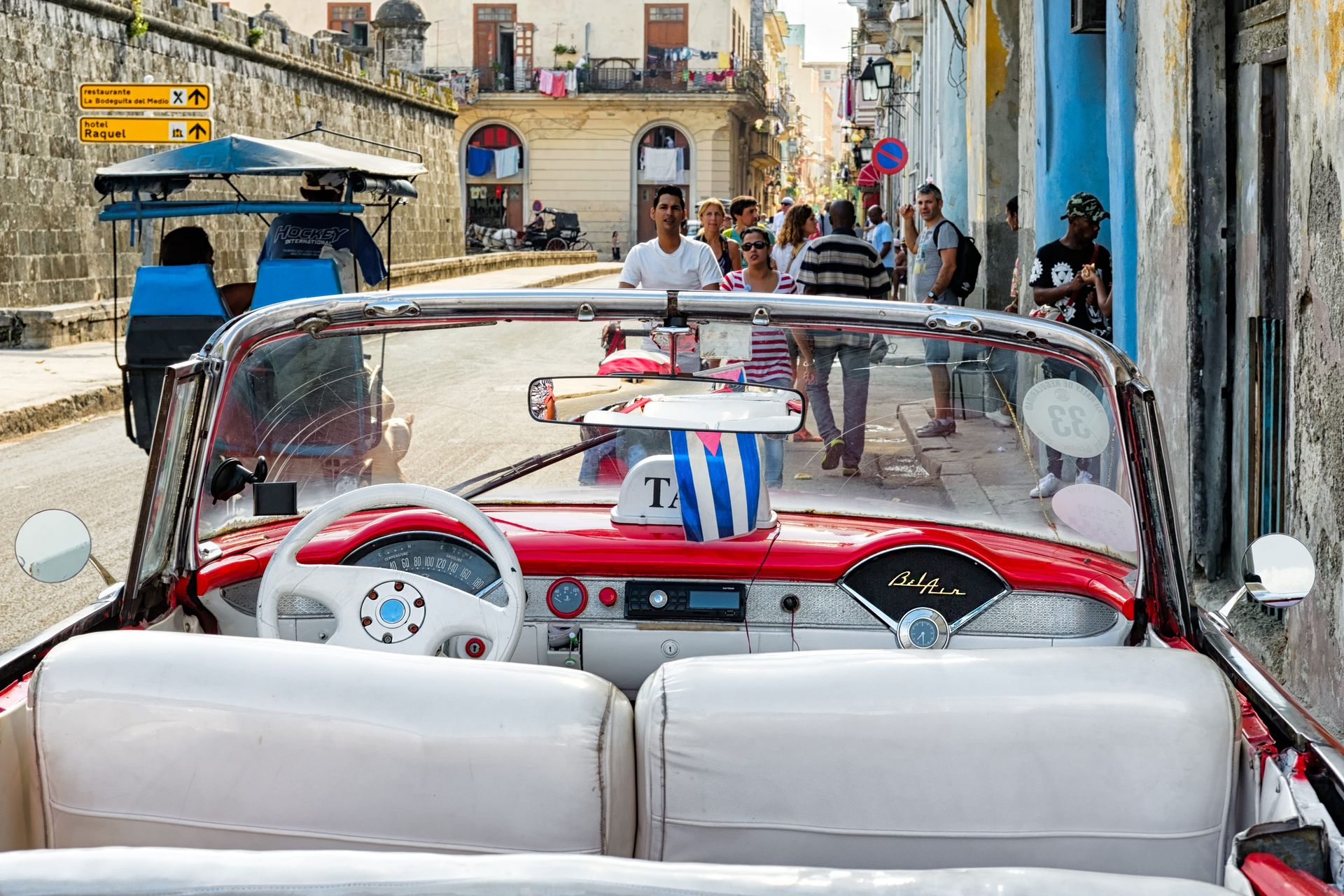 Classic red-and-white convertible driving through a narrow old stone street with pedestrians nearby